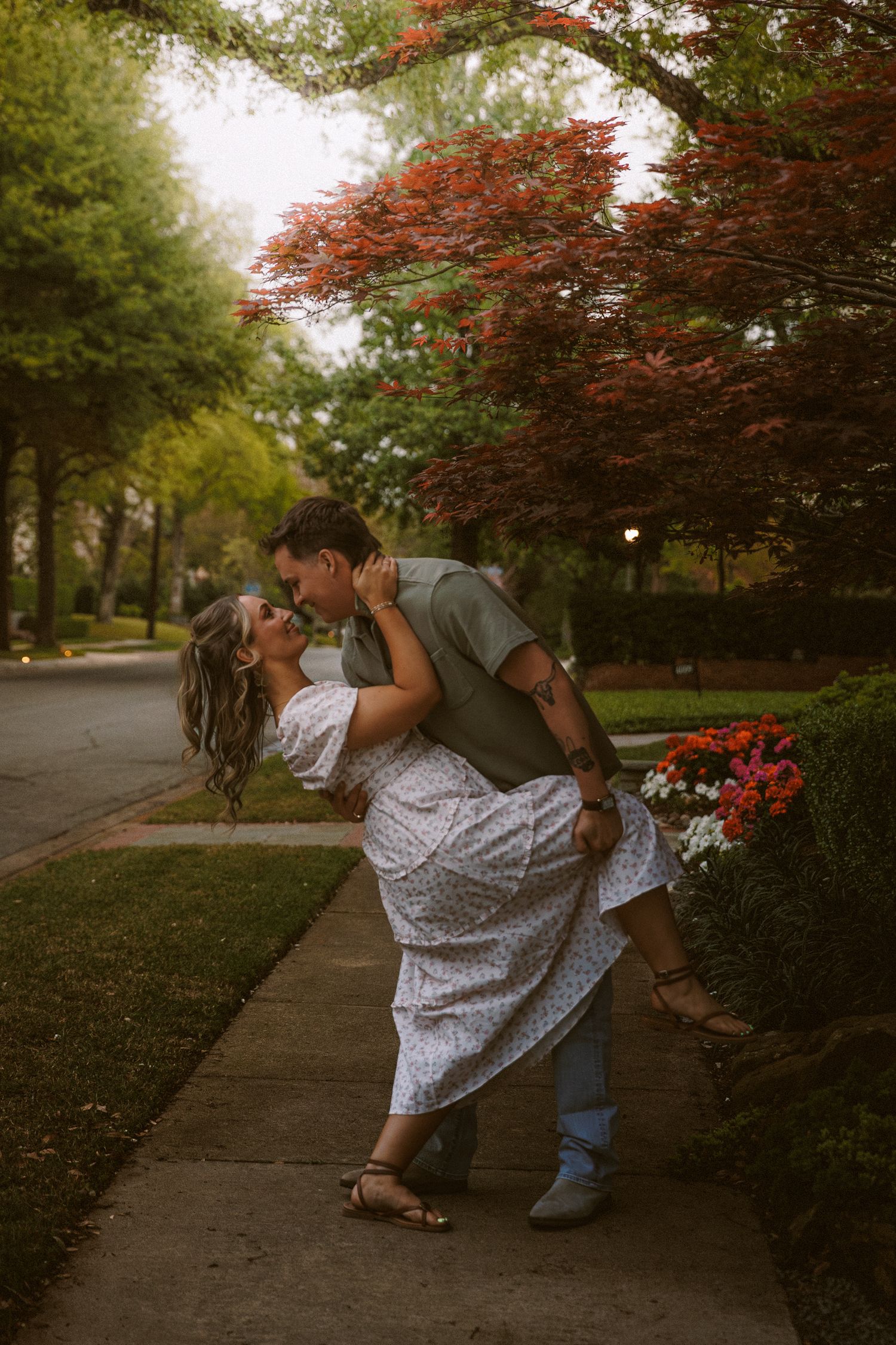 A couple shares a passionate dip kiss on a tree-lined sidewalk with colorful flowers.