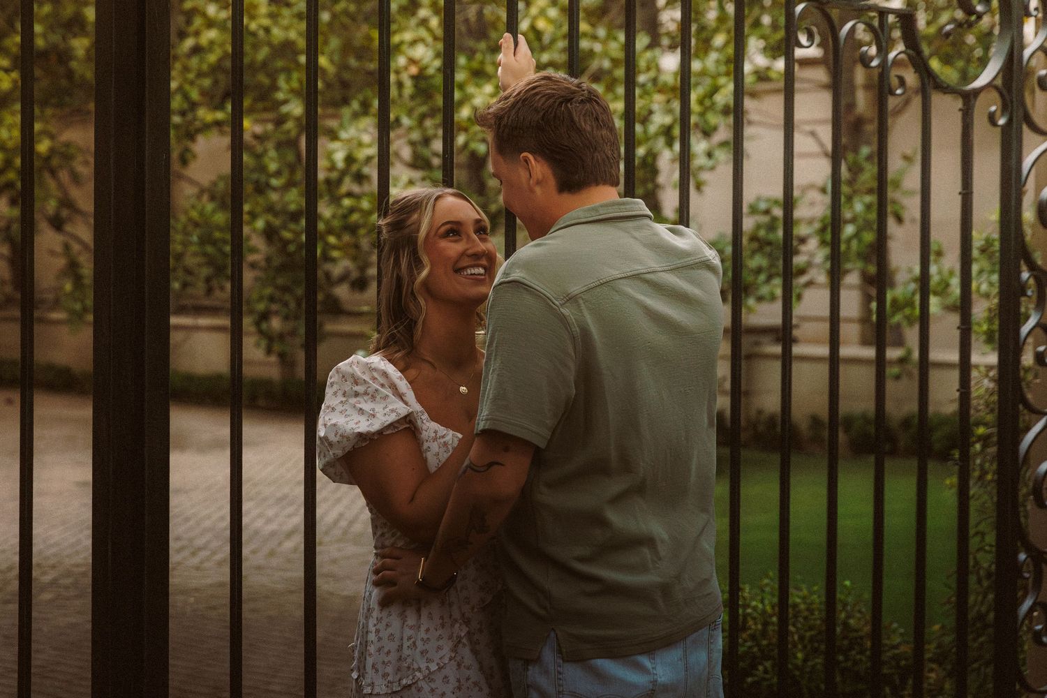 Smiling couple embracing by ornate iron gates surrounded by lush green foliage.
