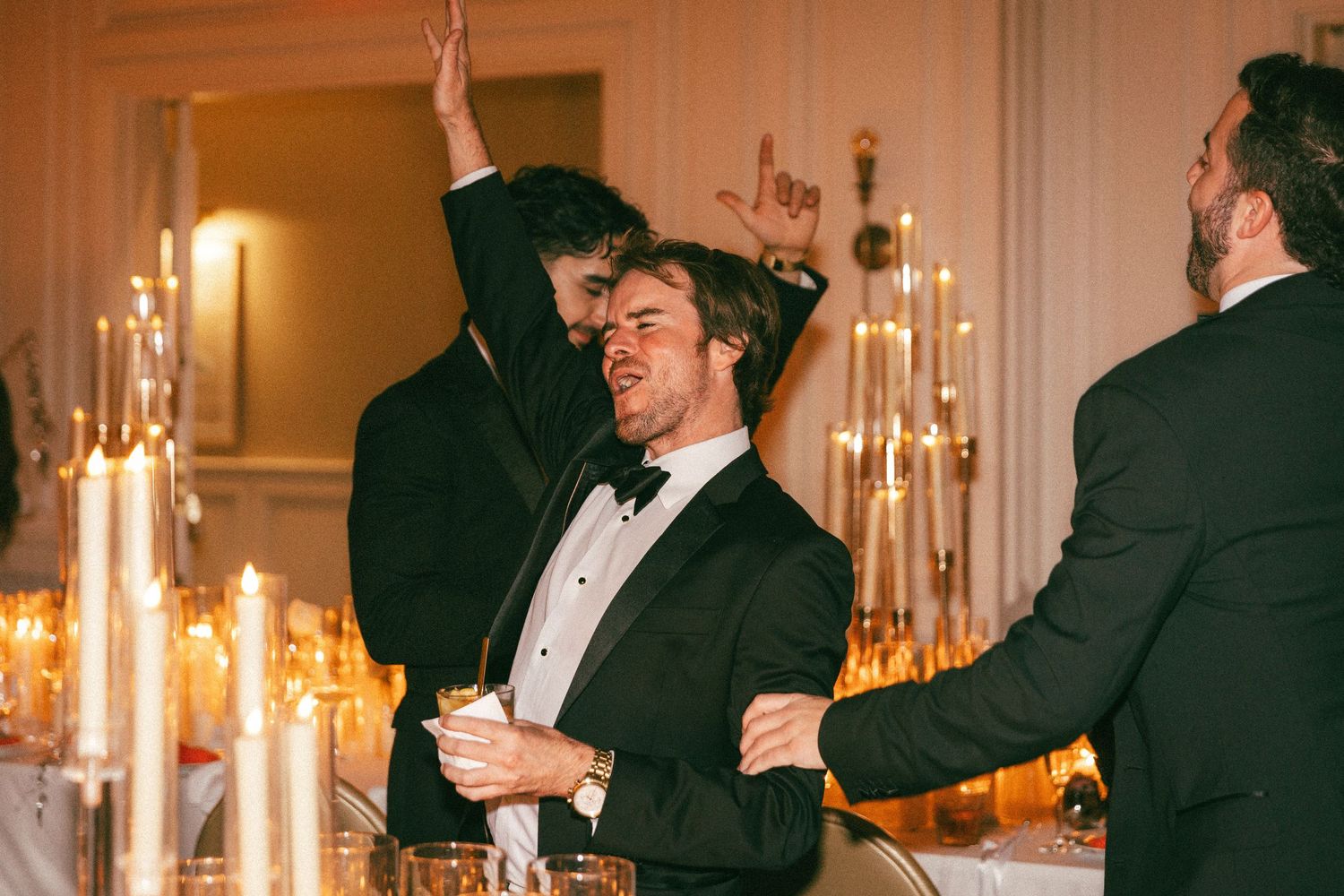 Guests in formal attire celebrate and dance near golden candelabras at a wedding reception.