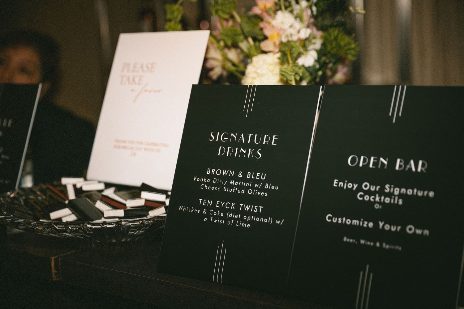 Elegant black and gold wedding bar signage displaying signature cocktails and open bar menu with floral arrangement.
