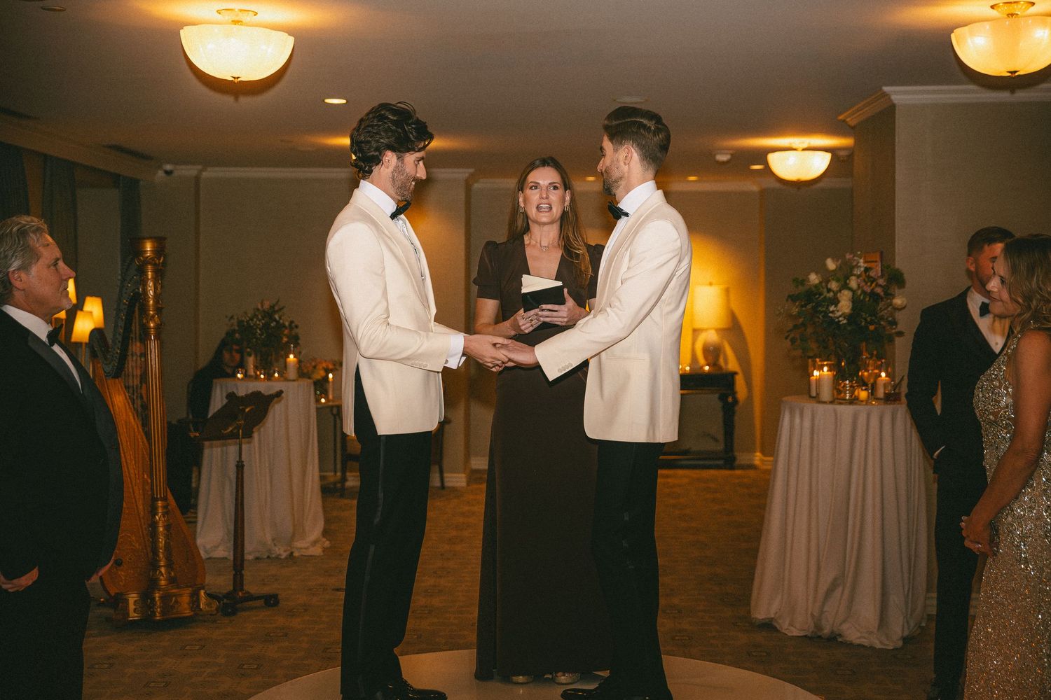 Two grooms in ivory tuxedos holding hands during an intimate wedding ceremony officiated by a woman in black.