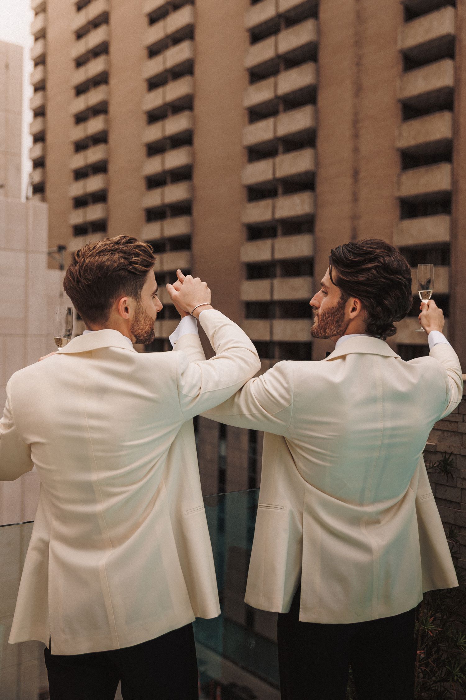 Two grooms in white tuxedo jackets toast with champagne on a rooftop with a city building backdrop.