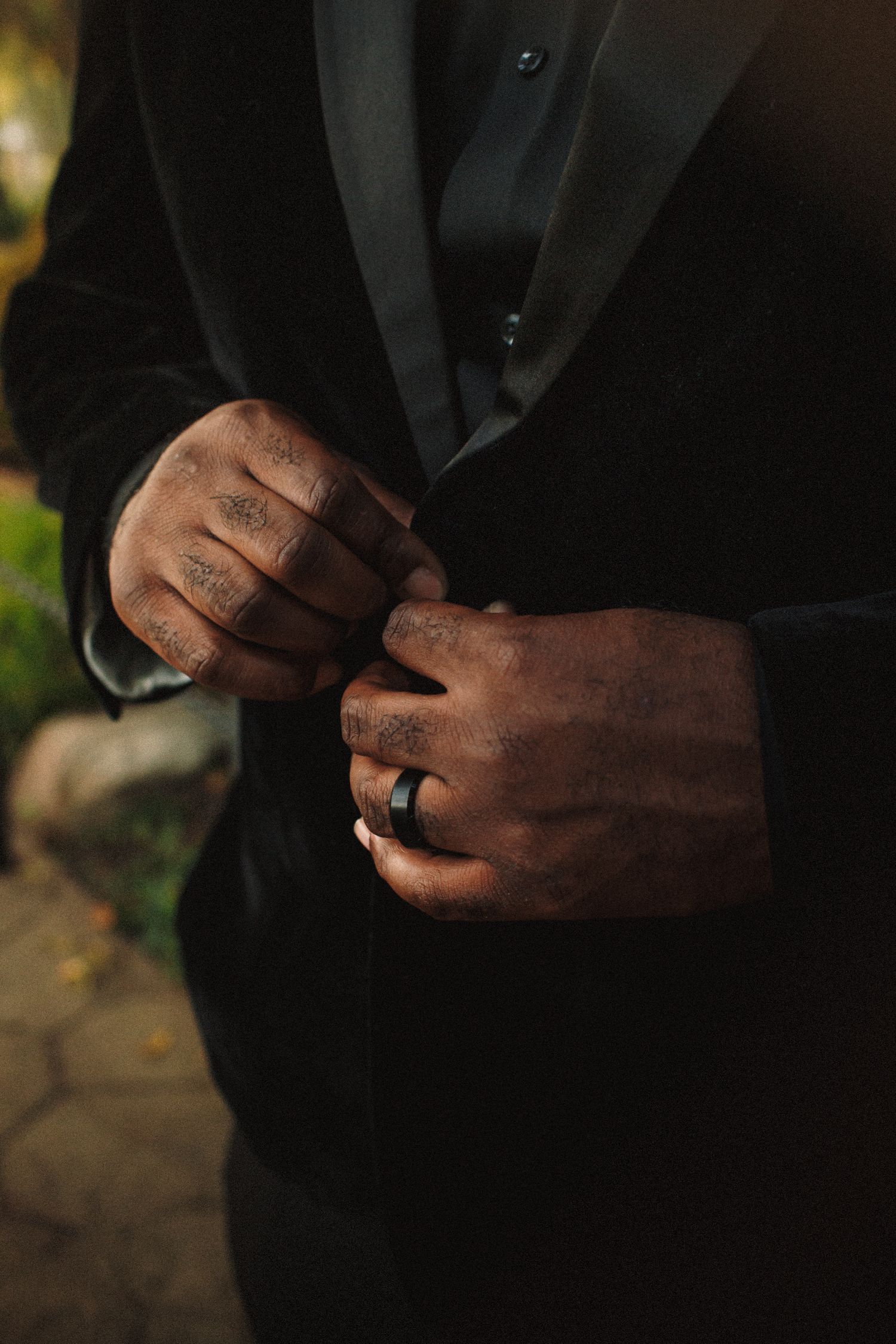 Close-up of groom adjusting black velvet tuxedo jacket with tattooed hands and black wedding band.