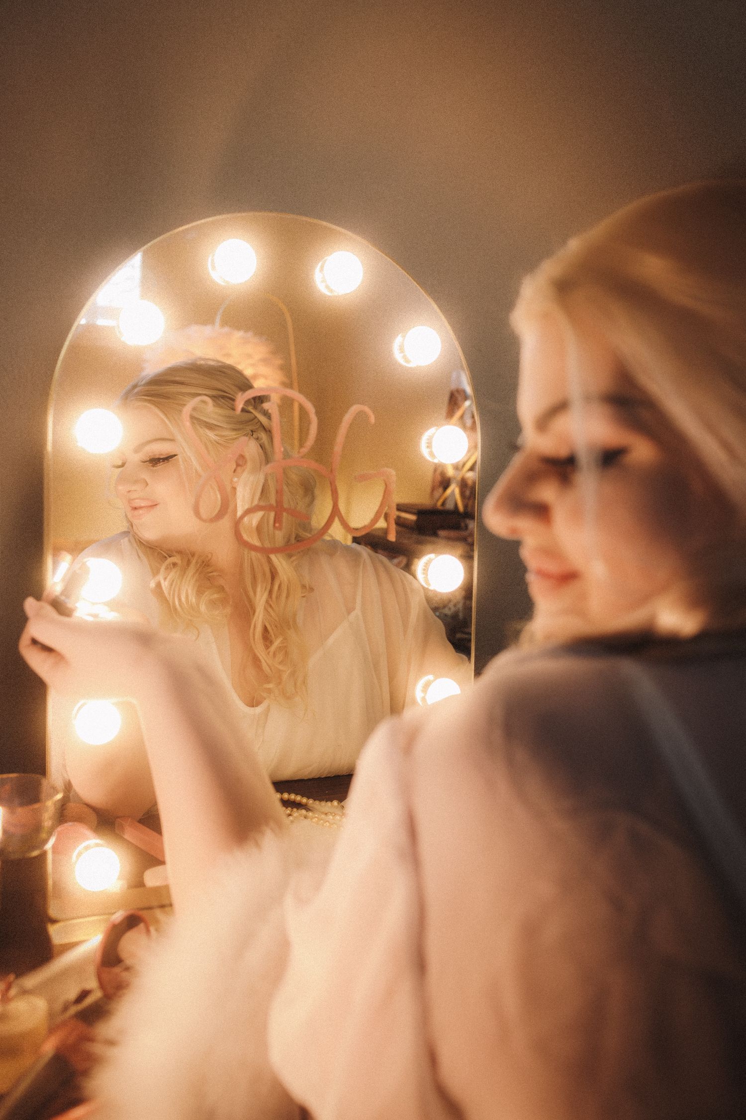 Blonde bride gazing into illuminated Hollywood vanity mirror with warm glowing bulbs reflection.