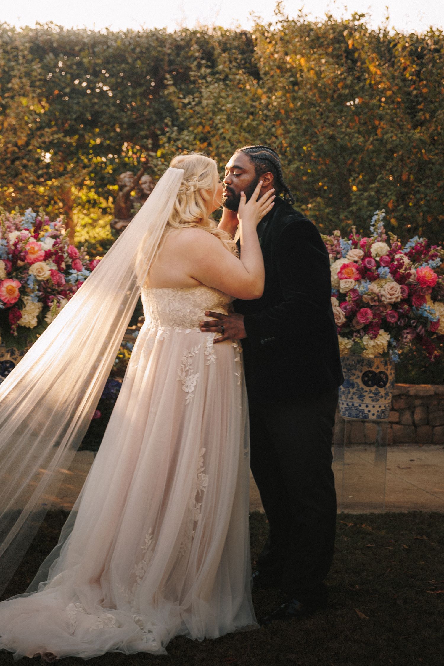 Bride and groom share their first kiss surrounded by colorful floral arrangements at an outdoor wedding ceremony.