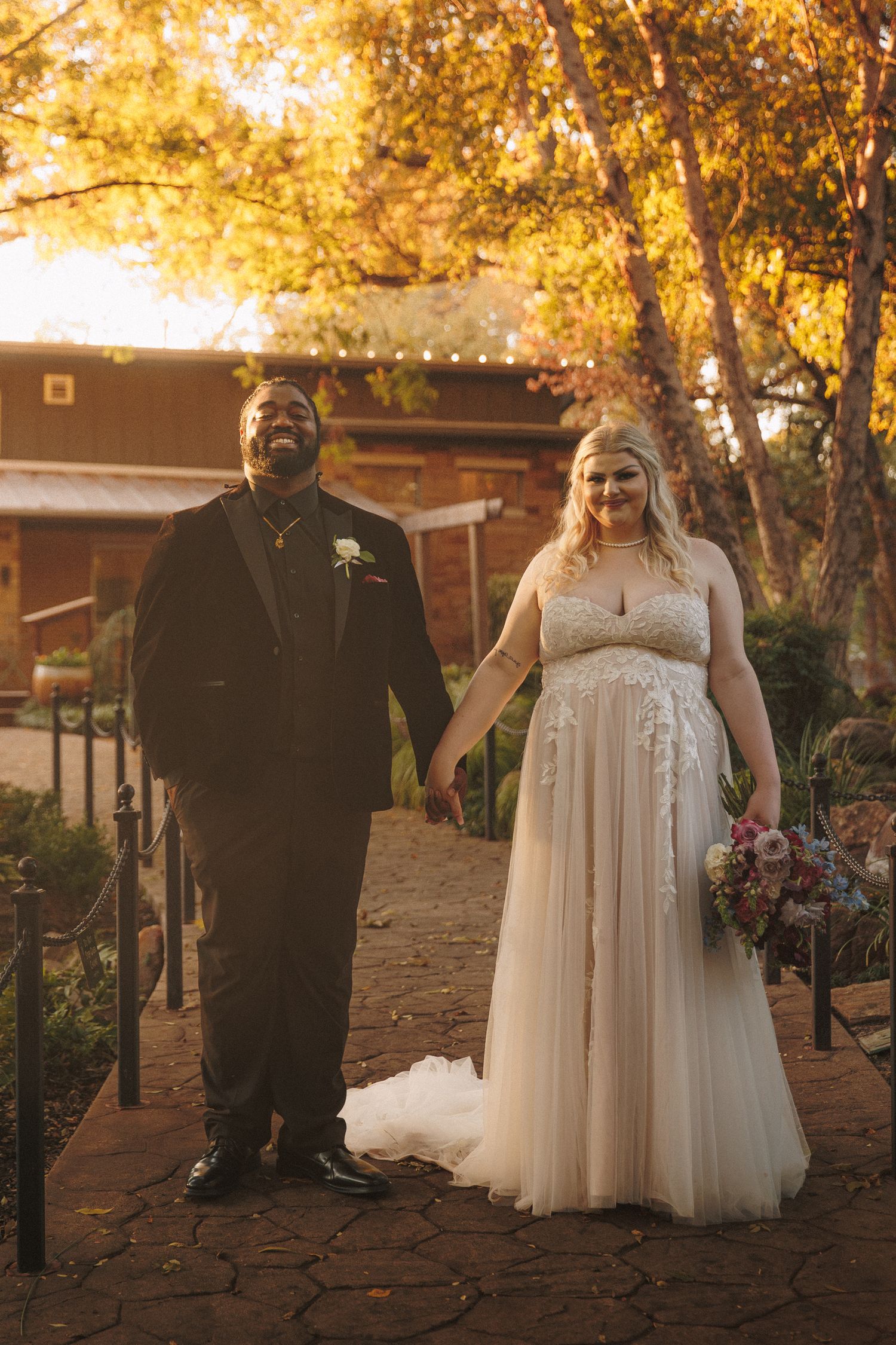 Smiling interracial couple holds hands in wedding attire on brick path with golden autumn trees.