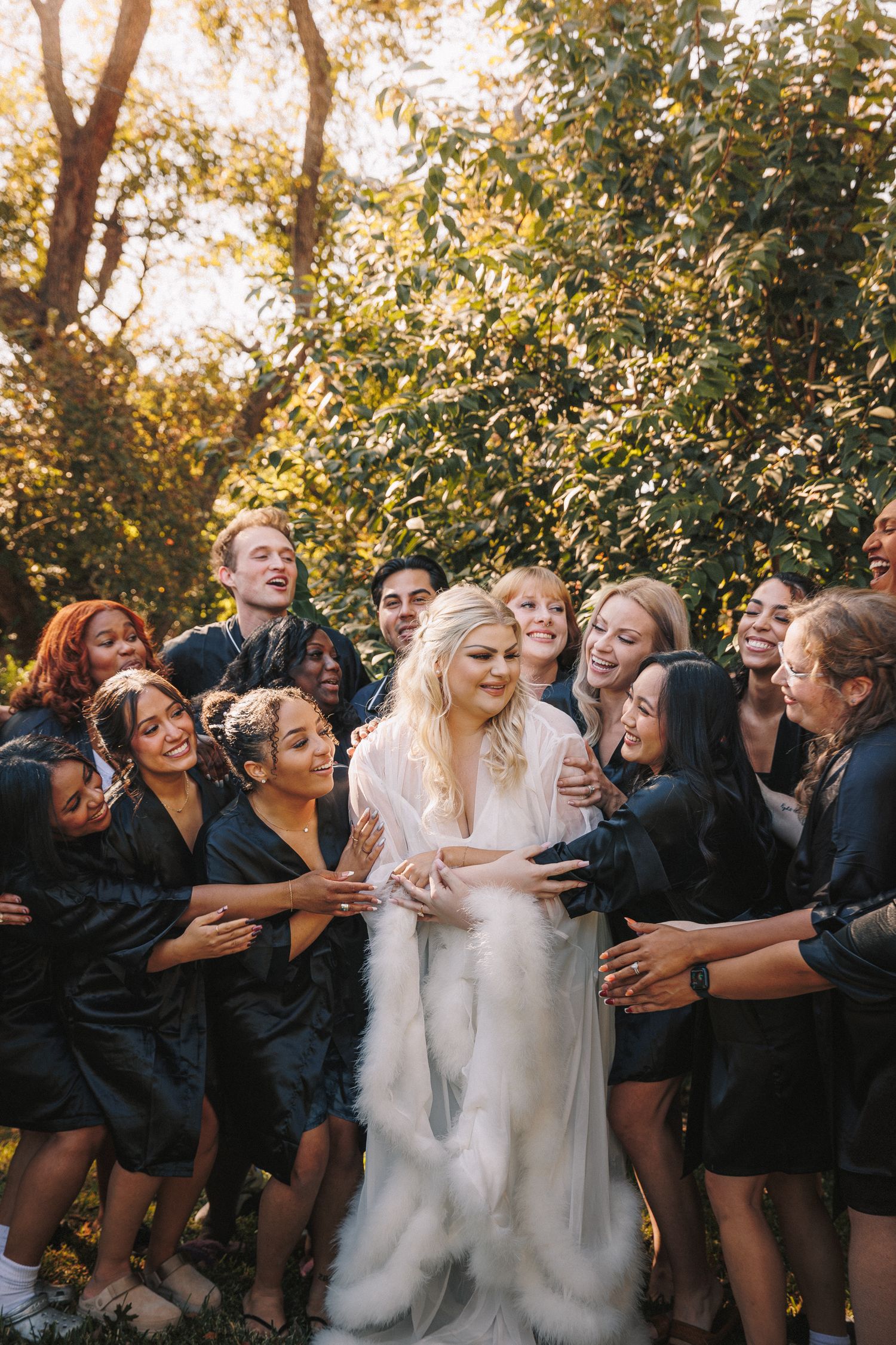 Bride in white gown and fur wrap surrounded by joyful wedding party outdoors in golden light.