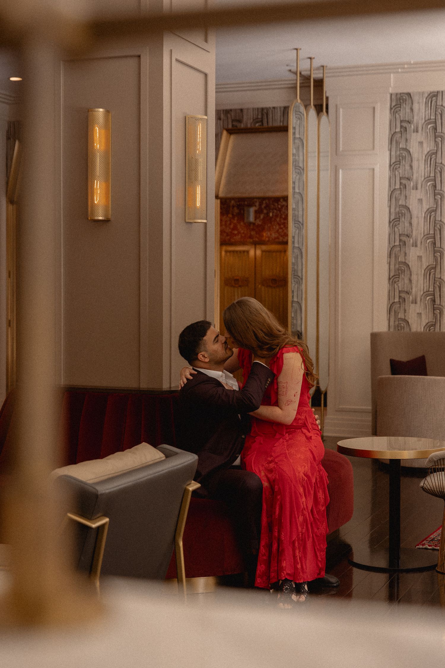 Couple sharing a romantic kiss in an elegant hotel lounge, woman in red dress seated on man's lap.