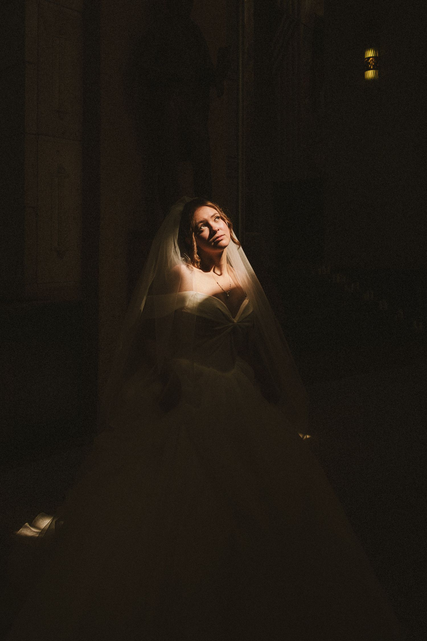 Bride in white ball gown and veil illuminated by dramatic spotlight in dark hall.