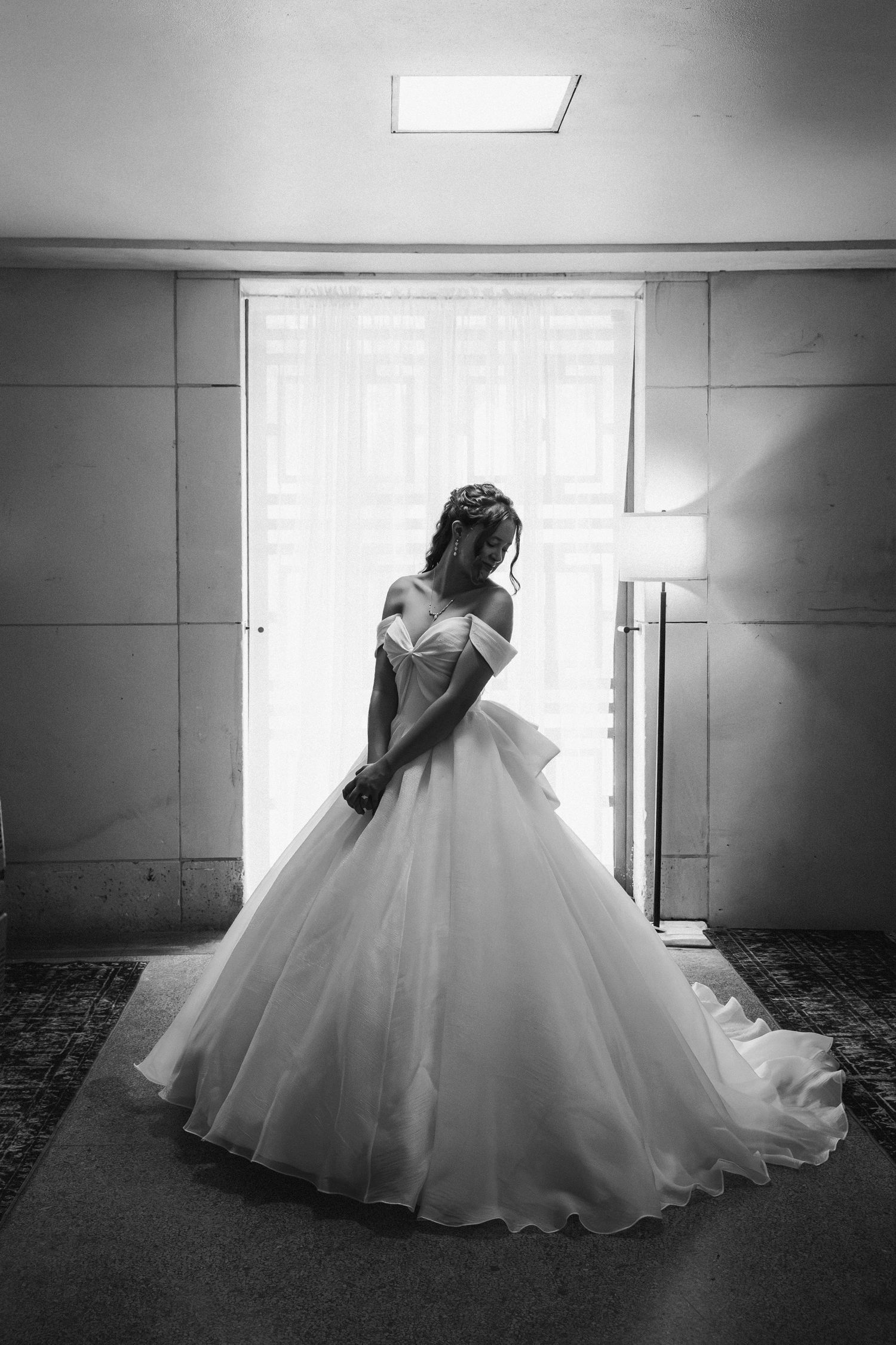 Bride in off-shoulder ball gown silhouetted against bright window in black and white.