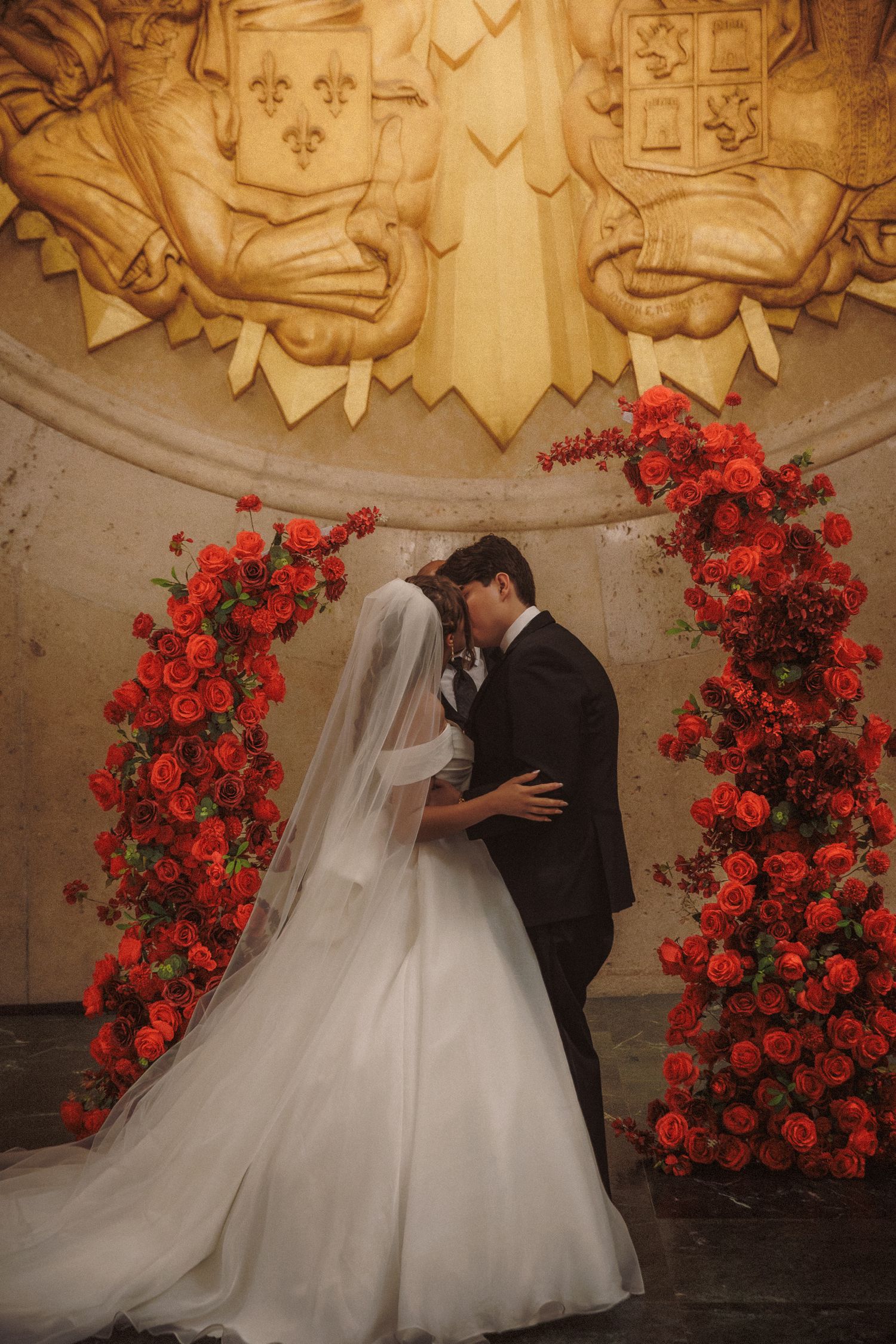 Bride and groom sharing first kiss under stunning red rose arch with gold crest backdrop.