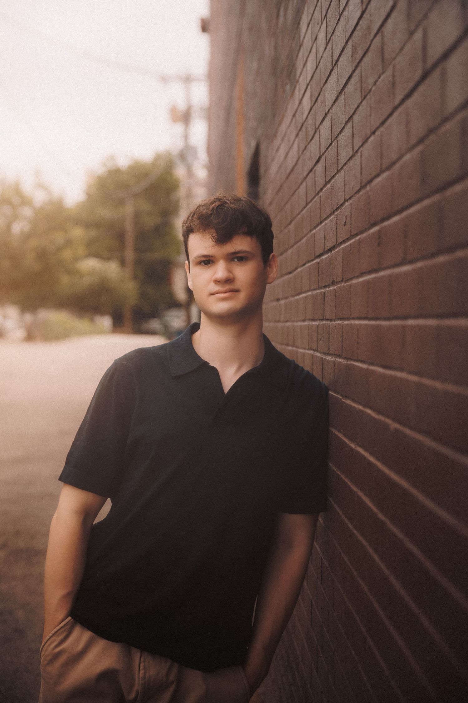 A person in black attire poses against a brick wall in natural golden-hour lighting.