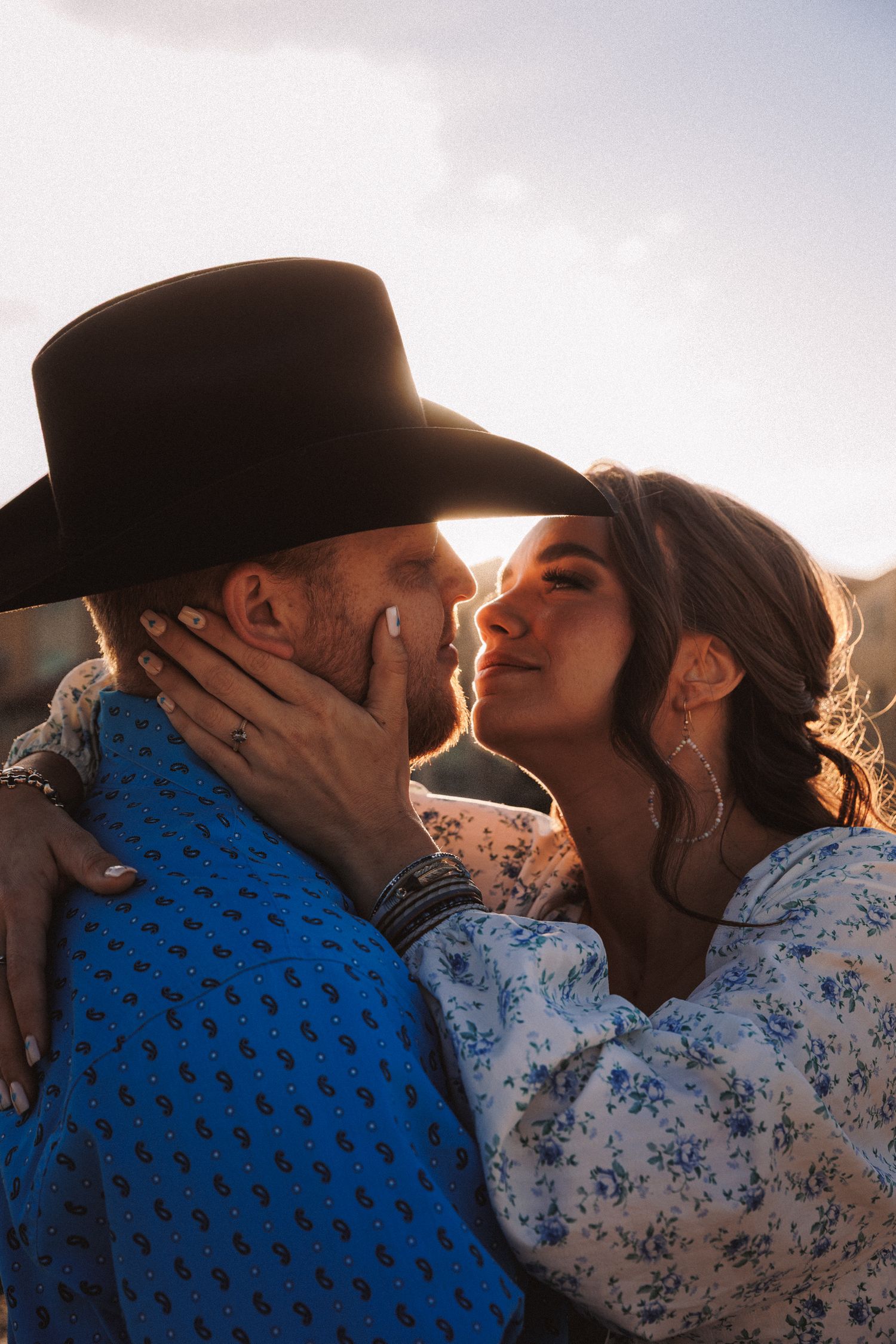 Romantic couple in western attire share an intimate moment at sunset, with her in floral dress and him in cowboy hat and blue shirt.