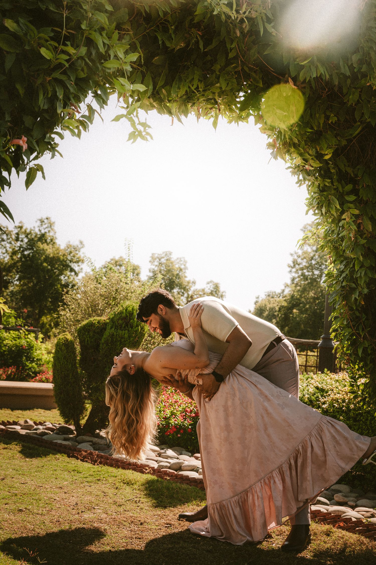 A couple performs a romantic dip pose in a garden setting during sunset.