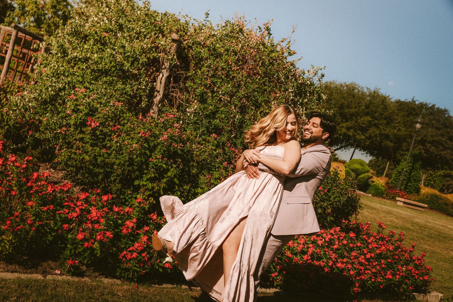 A couple embraces in a romantic dance pose among blooming red flowers and trees in a garden setting.