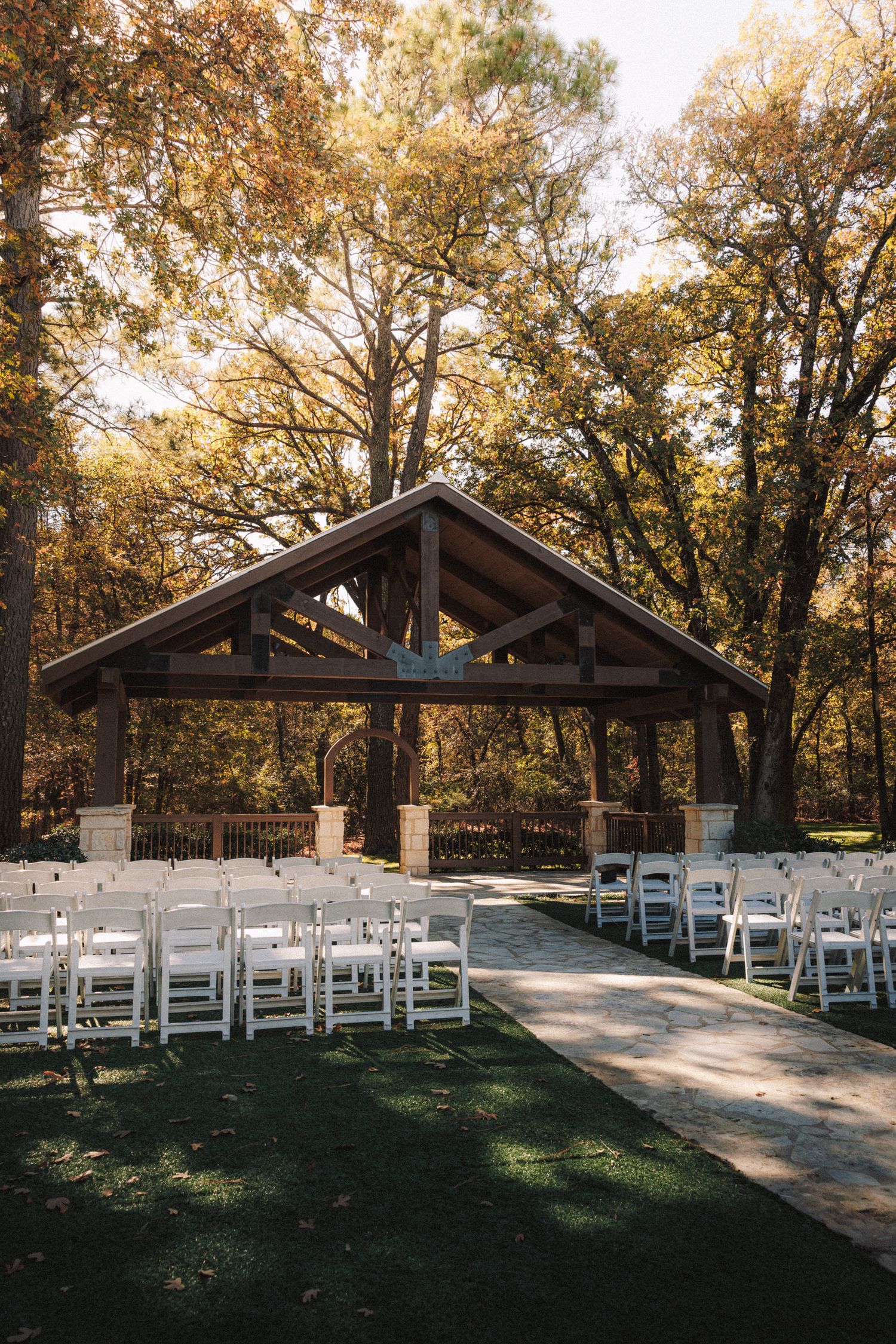 Outdoor wedding ceremony venue with white chairs and rustic wooden pavilion surrounded by autumn trees.