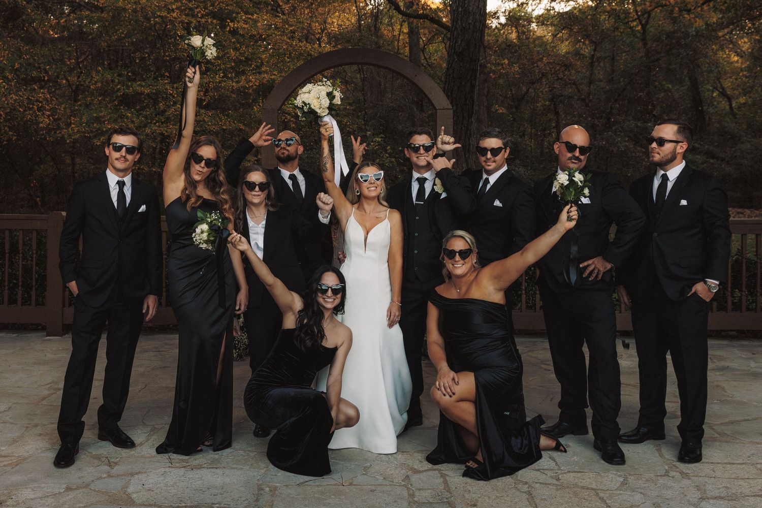 Fun wedding party photo with bride, groom, bridesmaids and groomsmen wearing sunglasses outdoors.