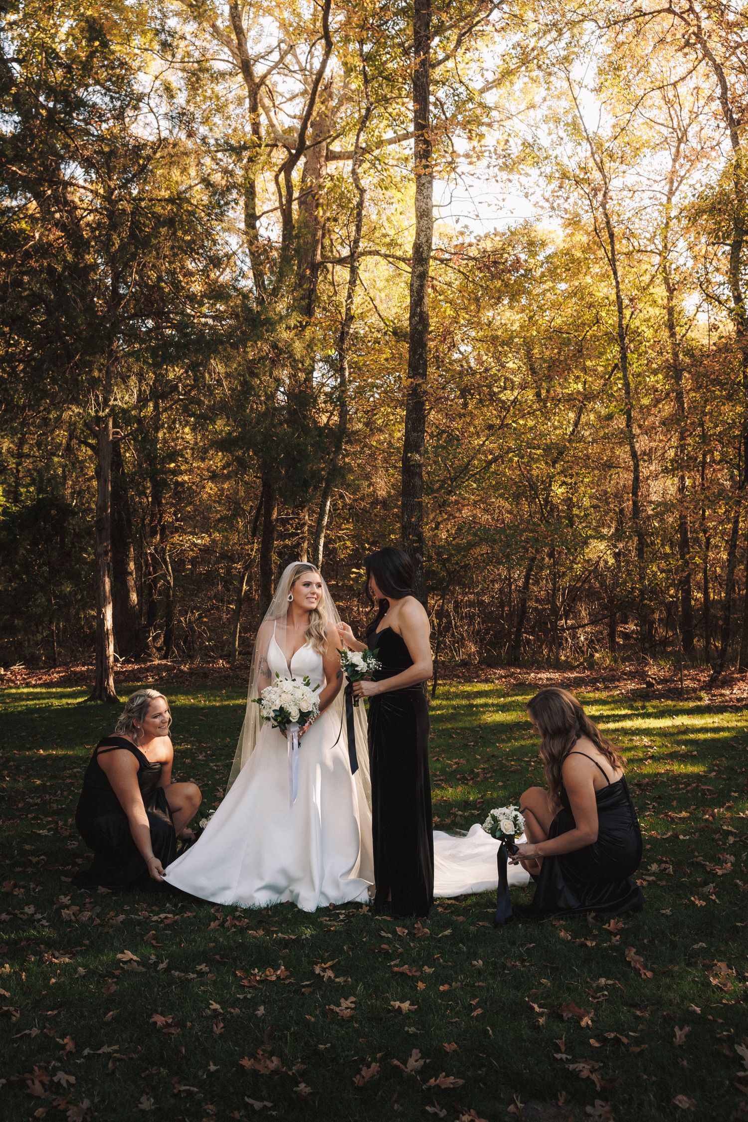 Bride in white gown with bridesmaids in black dresses adjusting her veil outdoors among autumn trees.