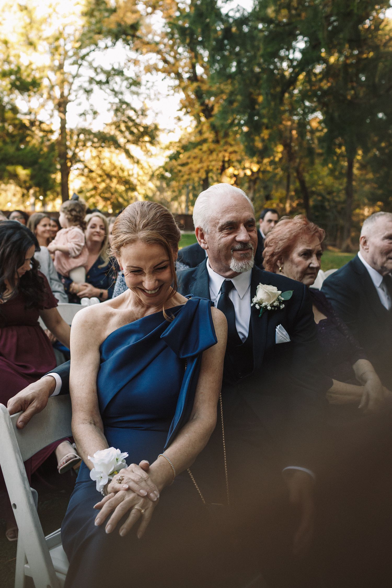 A smiling woman in a blue one-shoulder gown with a wrist corsage sits beside a man in a suit at an outdoor wedding ceremony.