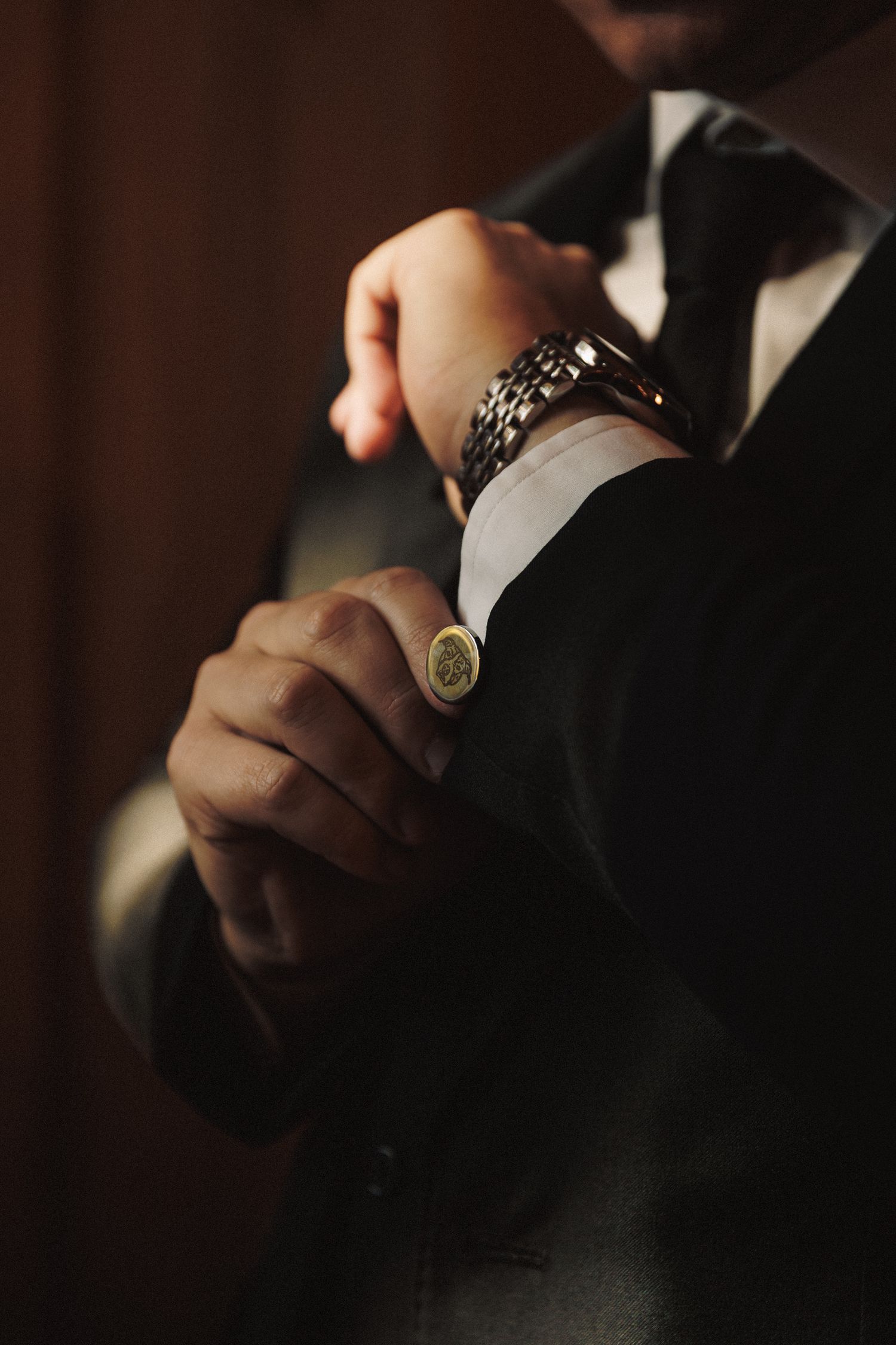 A groom in a dark suit adjusts his cufflinks, showcasing a gold signet ring and silver watch.