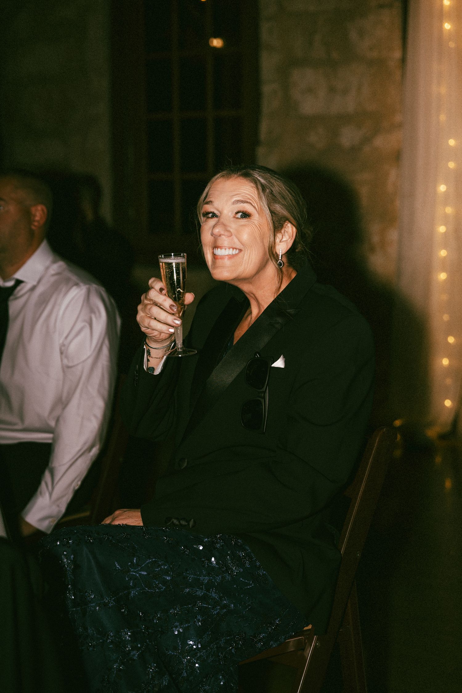 Elegantly dressed woman in black tuxedo jacket smiles while raising champagne glass at wedding reception.