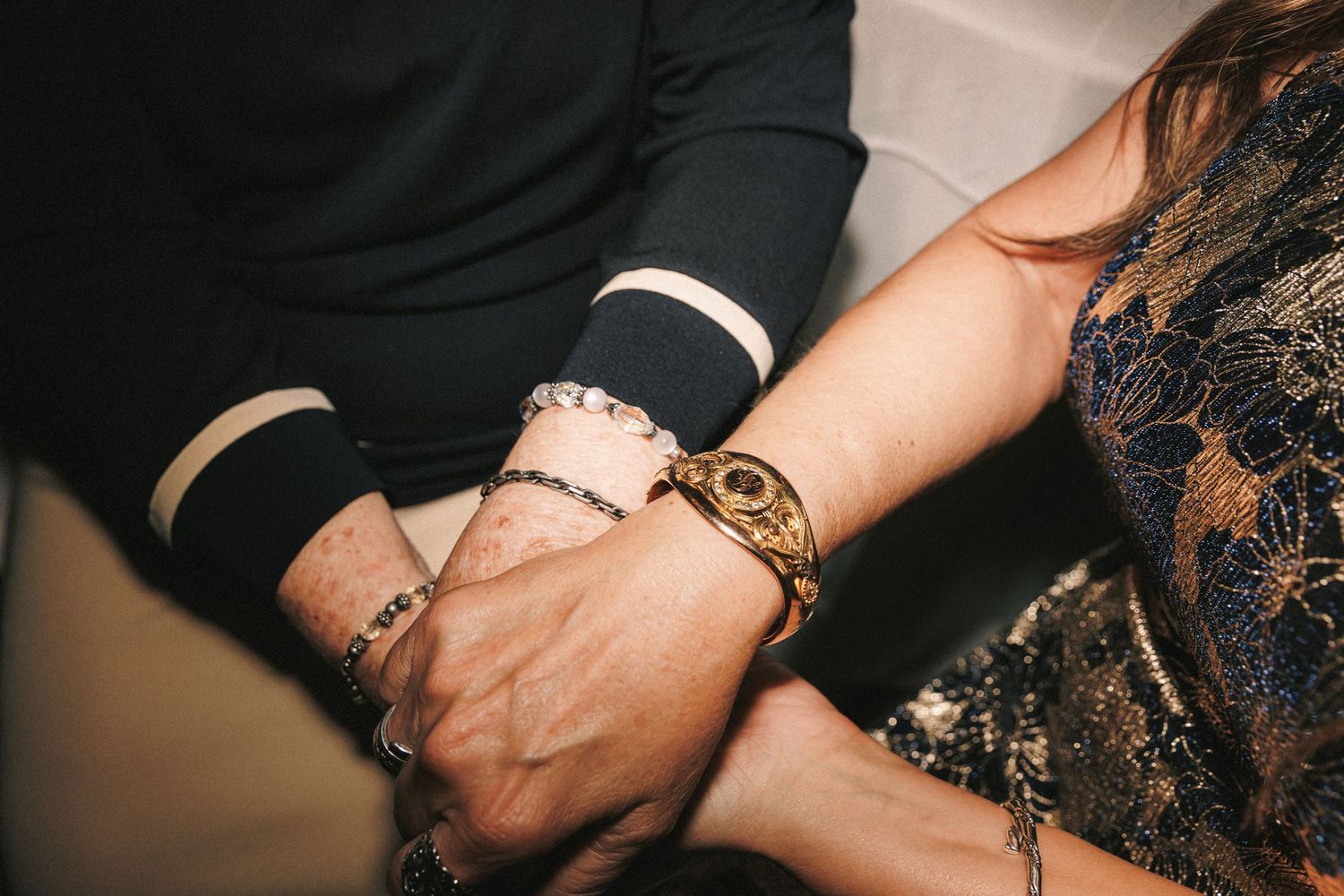 Close-up of two people holding hands wearing elegant bracelets and watches at a wedding reception.