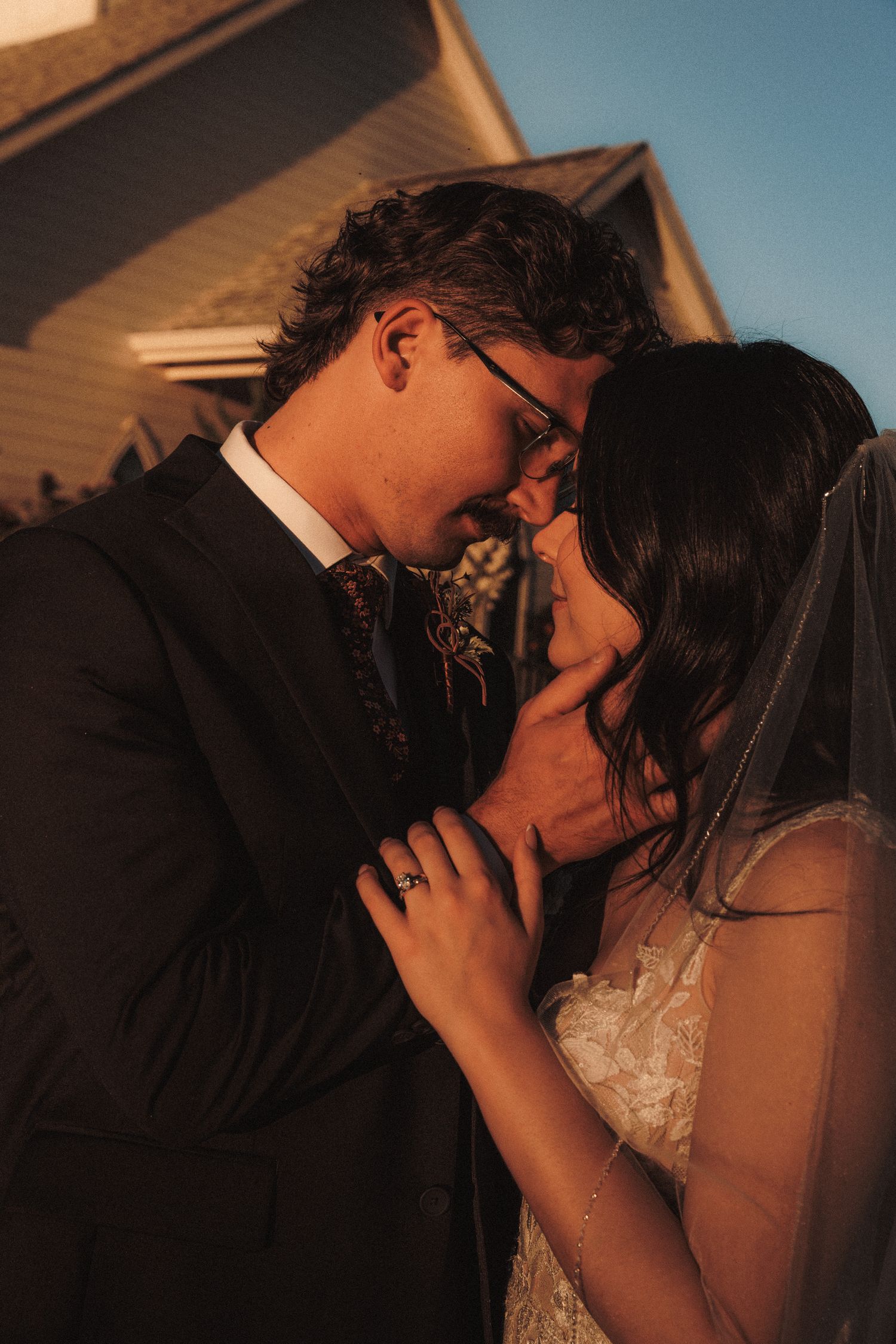 Bride and groom share a romantic forehead touch in warm golden hour light outside a chapel.