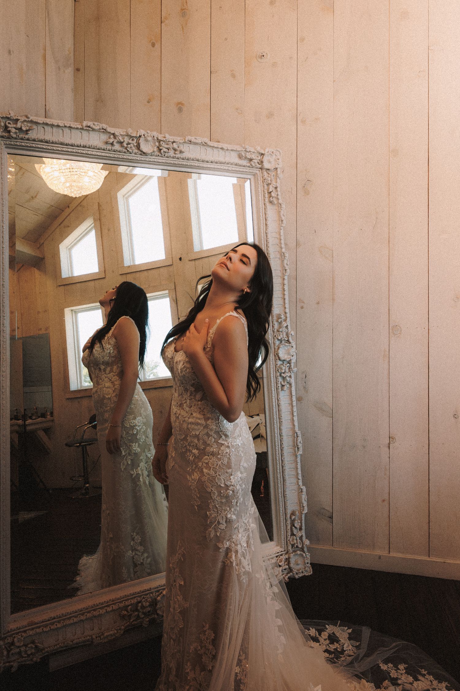 Bride in lace mermaid gown poses elegantly beside ornate white mirror reflecting rustic interior.