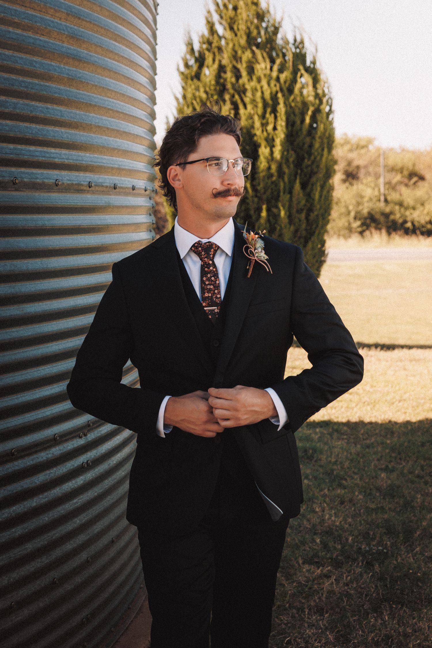 Groom in black suit with mustache and glasses adjusting jacket outdoors near rustic grain silo.