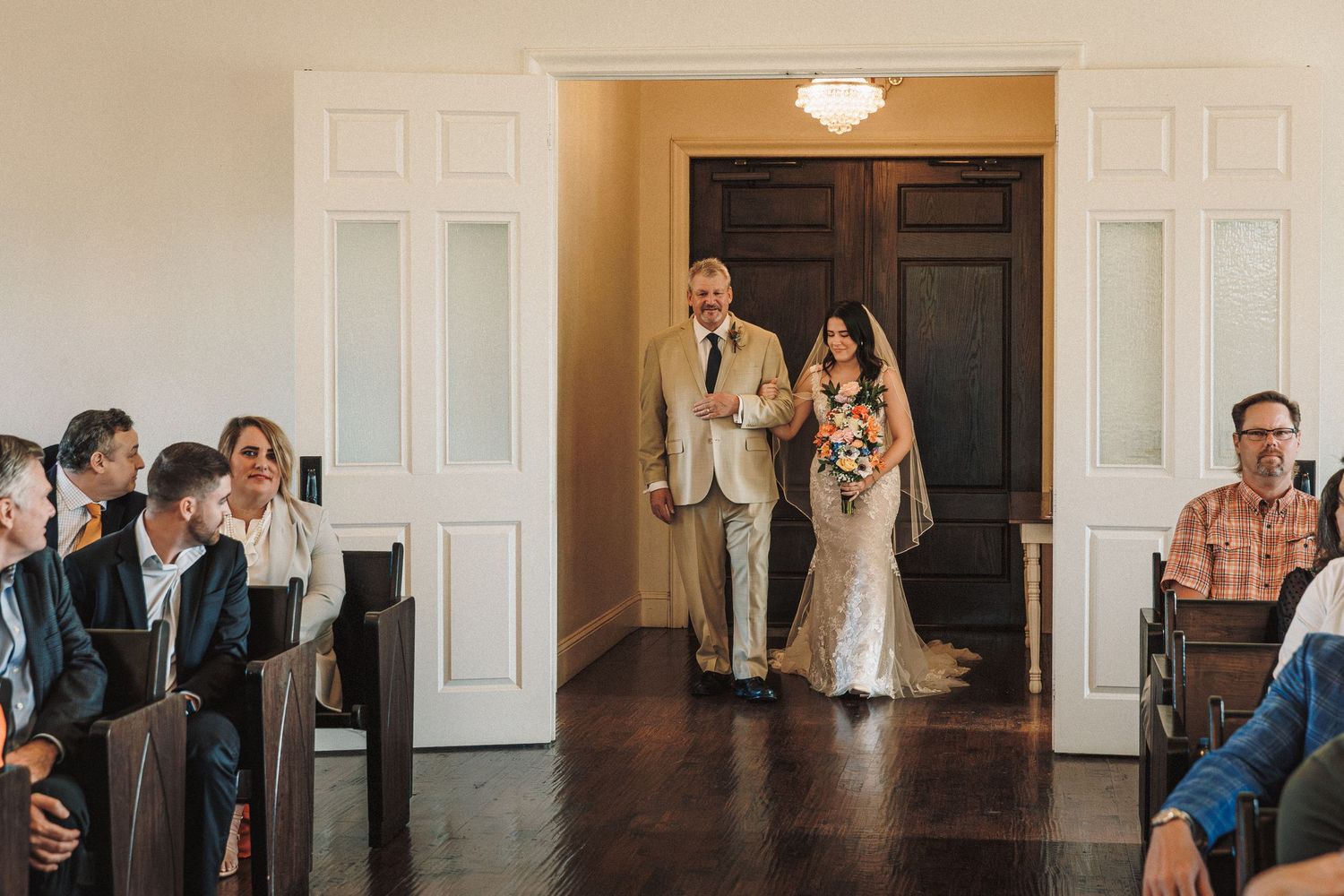 Bride in lace gown walks down chapel aisle with escort as seated wedding guests look on.