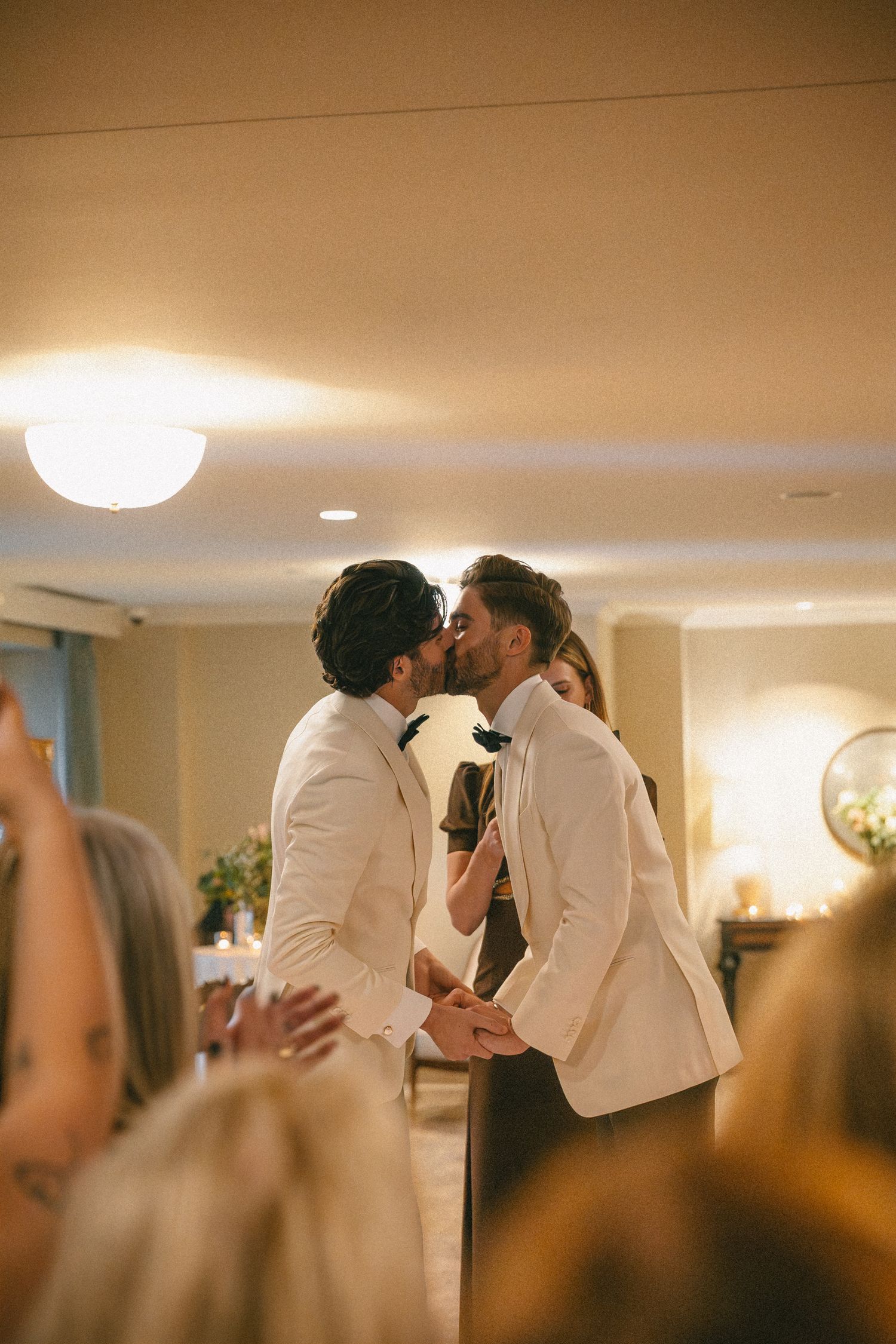 Two grooms in matching white tuxedos share a first kiss at their wedding reception.
