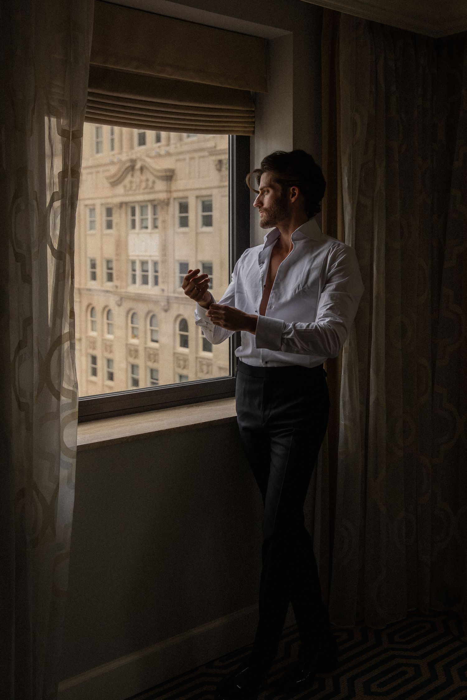 Elegant groom in white shirt gazing out hotel window at city buildings while getting dressed.