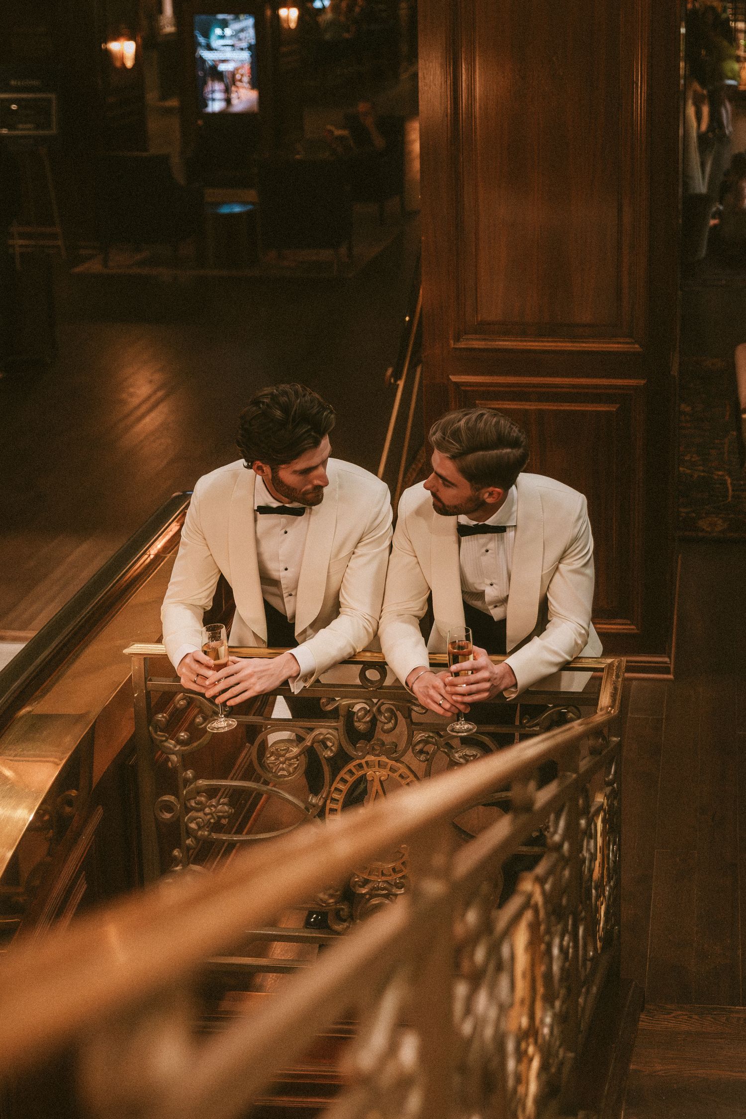 Two grooms in matching white tuxedos leaning on ornate gold railing sharing champagne together.