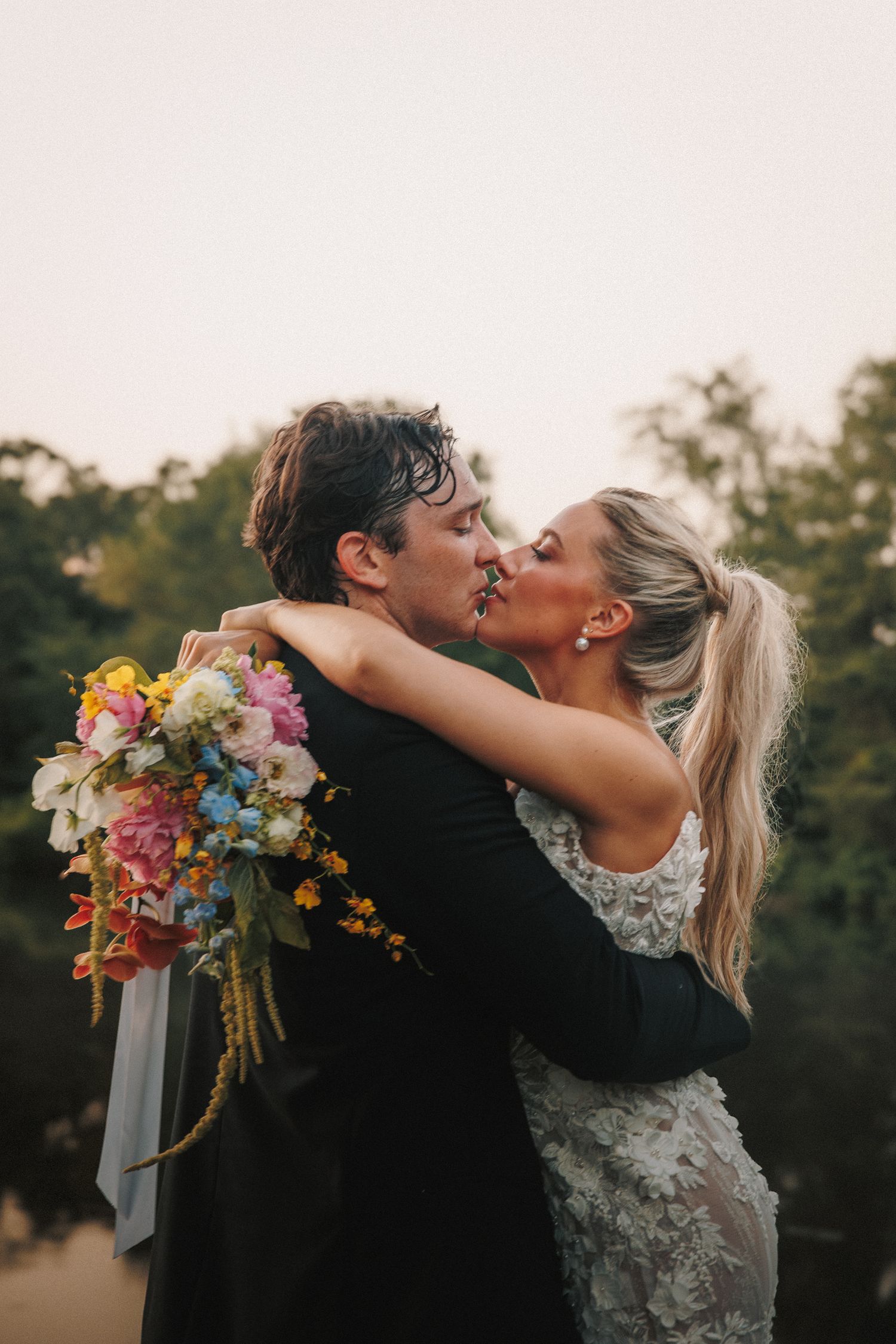 A couple embraces in a romantic kiss outdoors while bride holds a vibrant bouquet of colorful summer flowers.