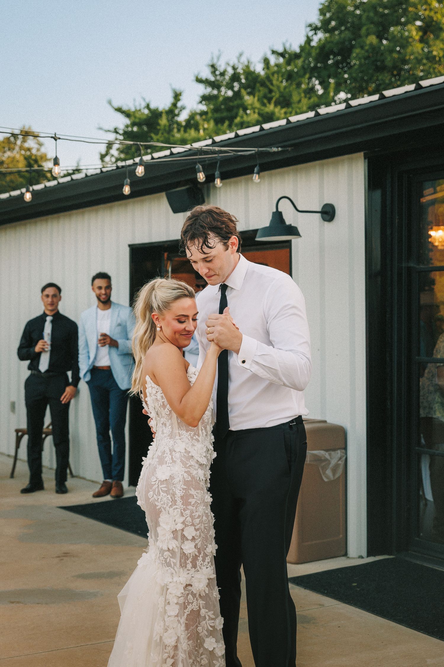 A couple dances together at an outdoor wedding venue with string lights and white buildings in the background.