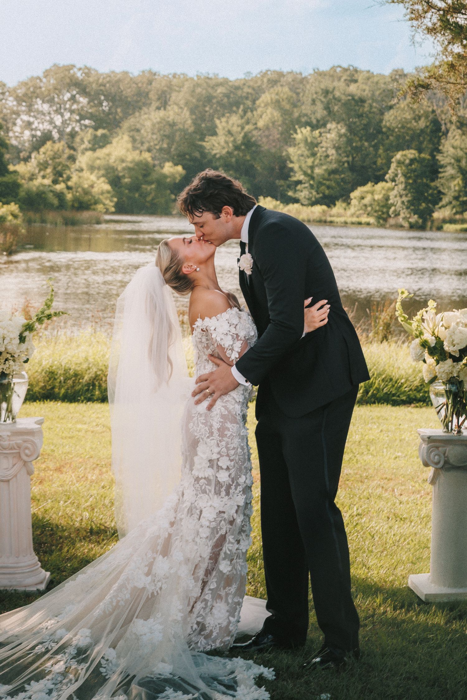 An outdoor wedding ceremony takes place near decorative white pillars beside a serene lake.