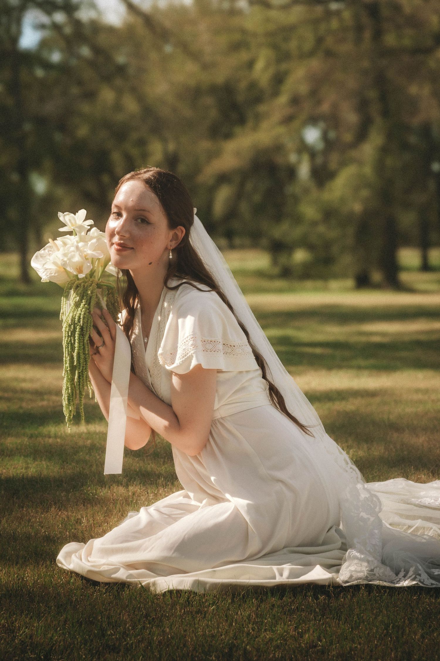 A bride in a white dress and veil lying in grass holding flowers in an outdoor garden setting.