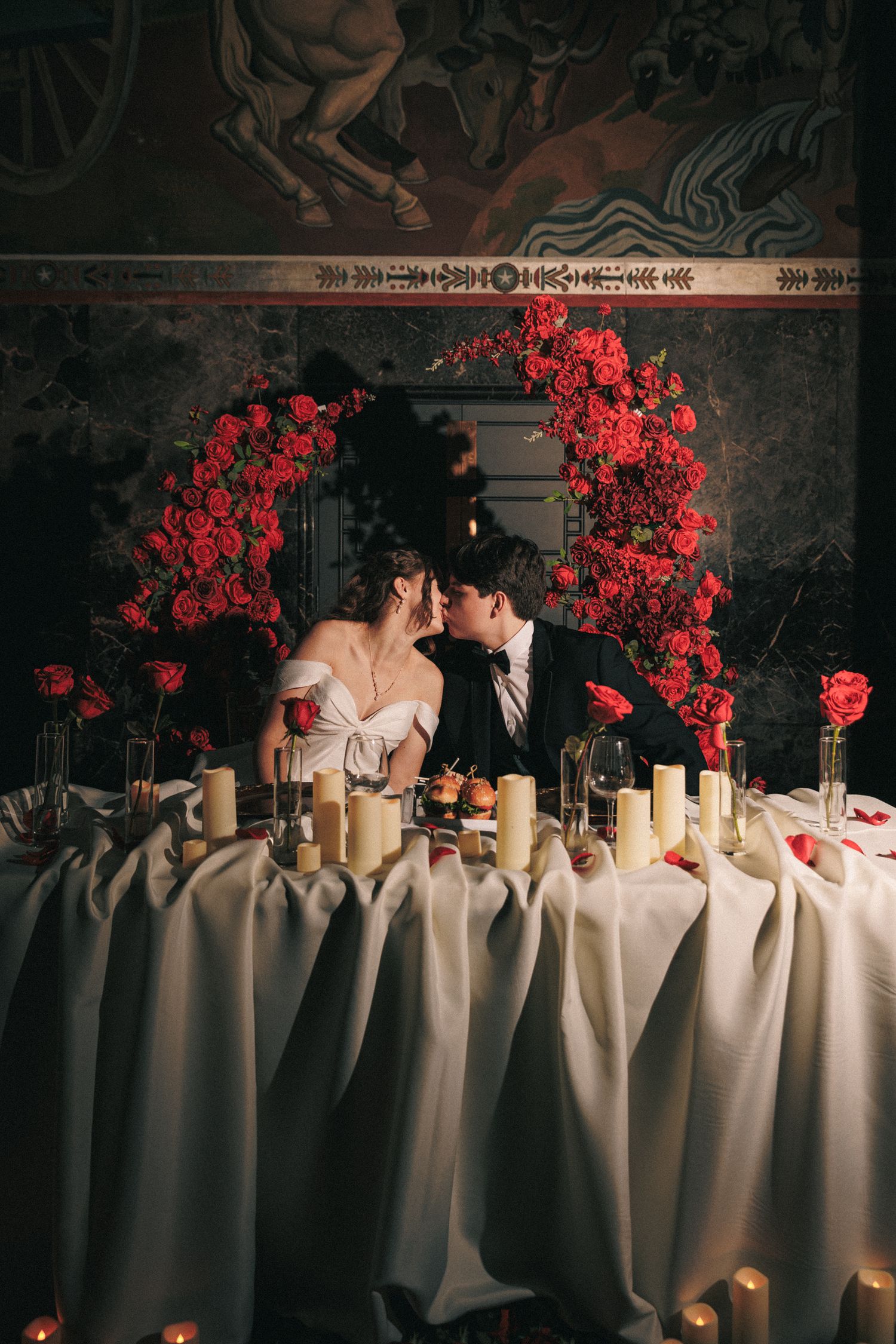 Newlyweds sharing intimate moment at candlelit sweetheart table with red rose arch behind.