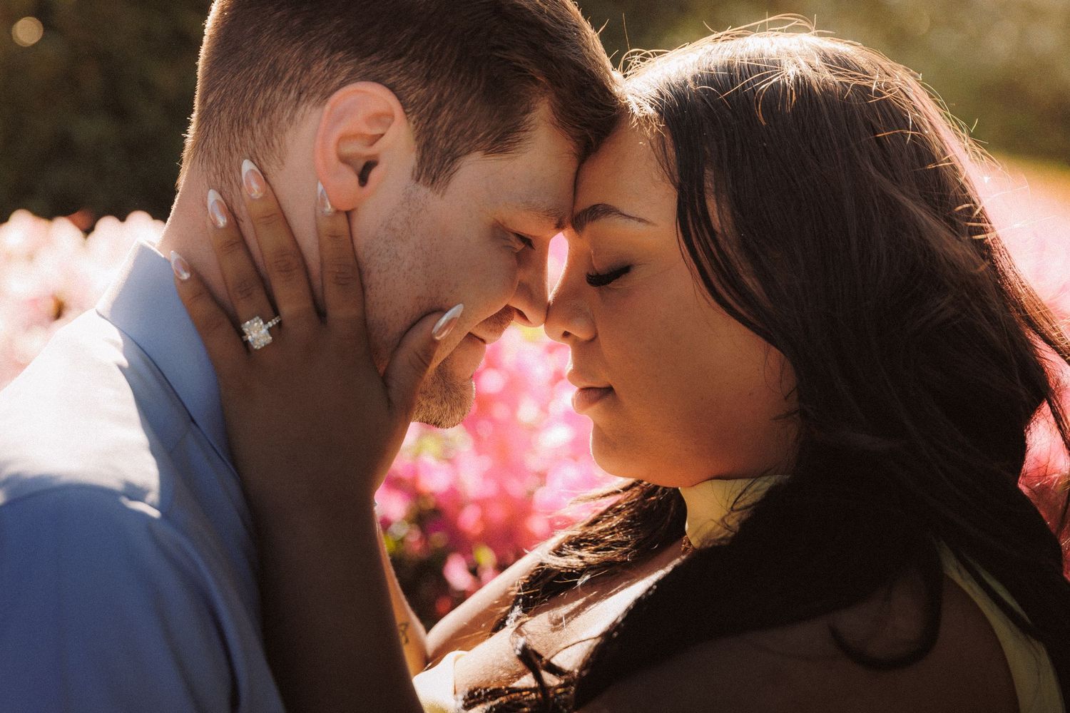 Romantic couple touching foreheads tenderly outdoors with pink blossoms in soft golden sunlight, woman wearing engagement ring.