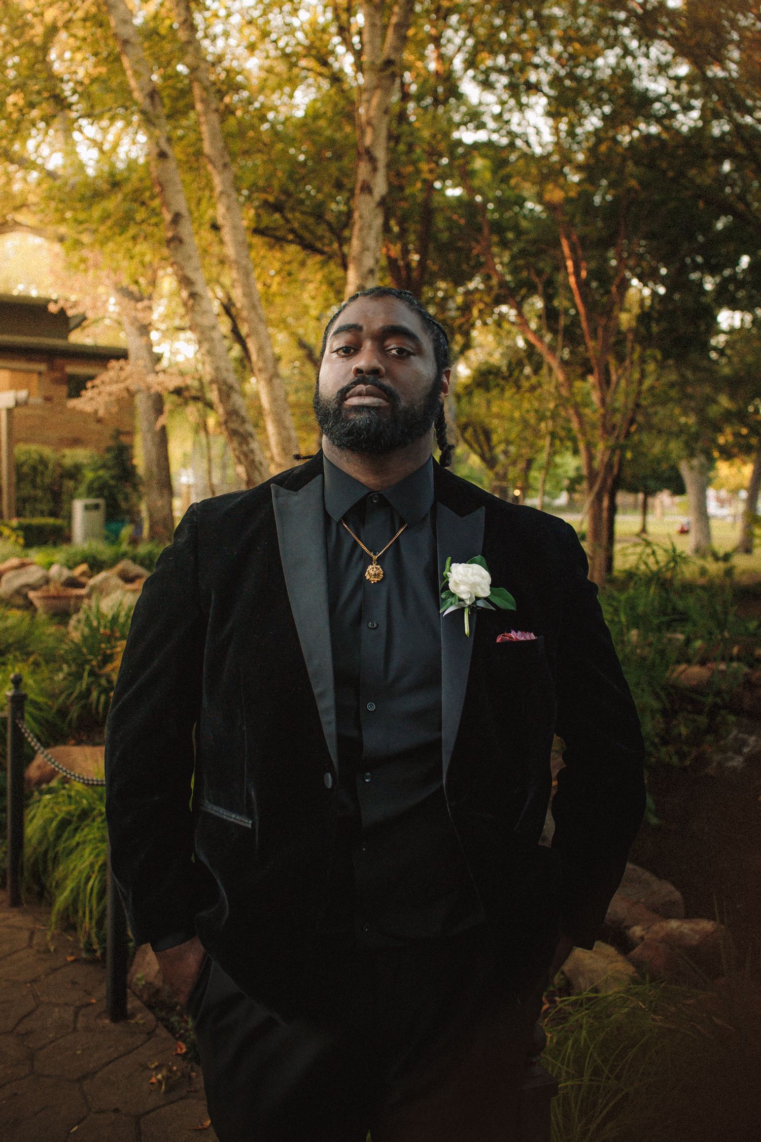 Groom in black velvet tuxedo with white boutonniere standing confidently in garden at sunset.