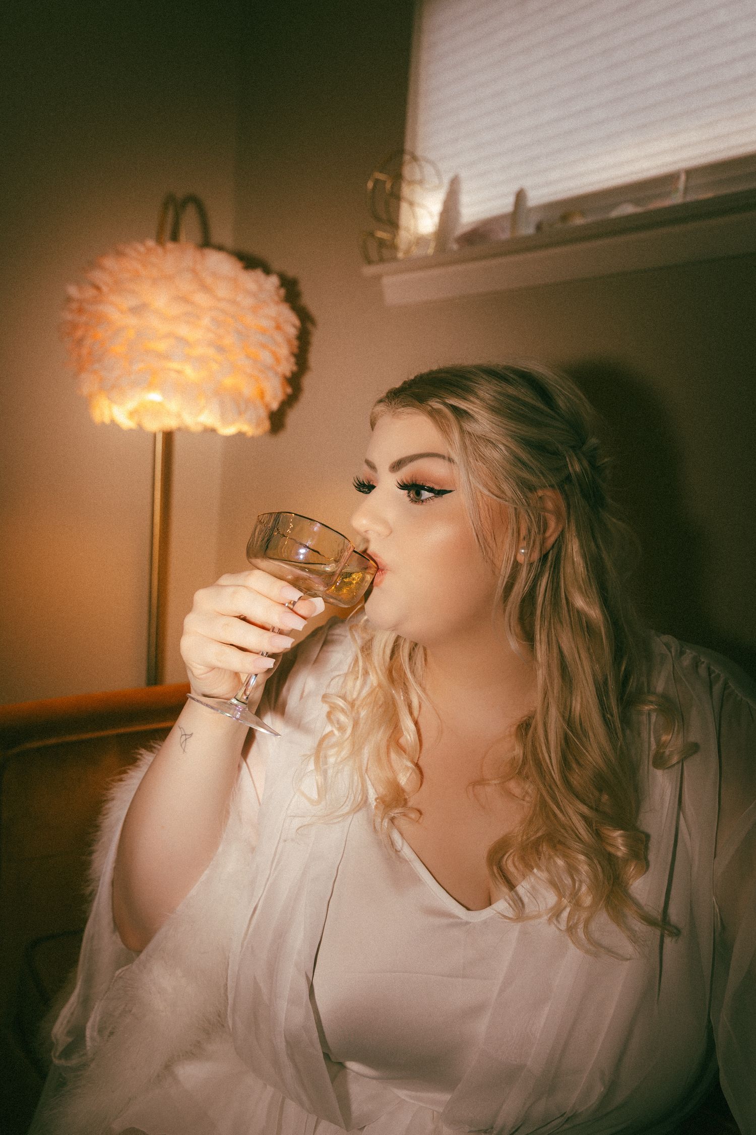 Bride in white robe sipping whiskey by warm lamp light during wedding morning preparations.