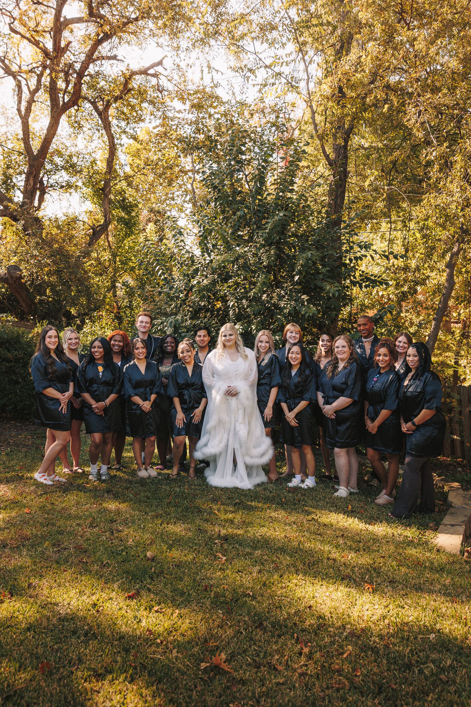 Bride in white gown poses with large bridal party in matching dark robes outdoors in autumn garden.