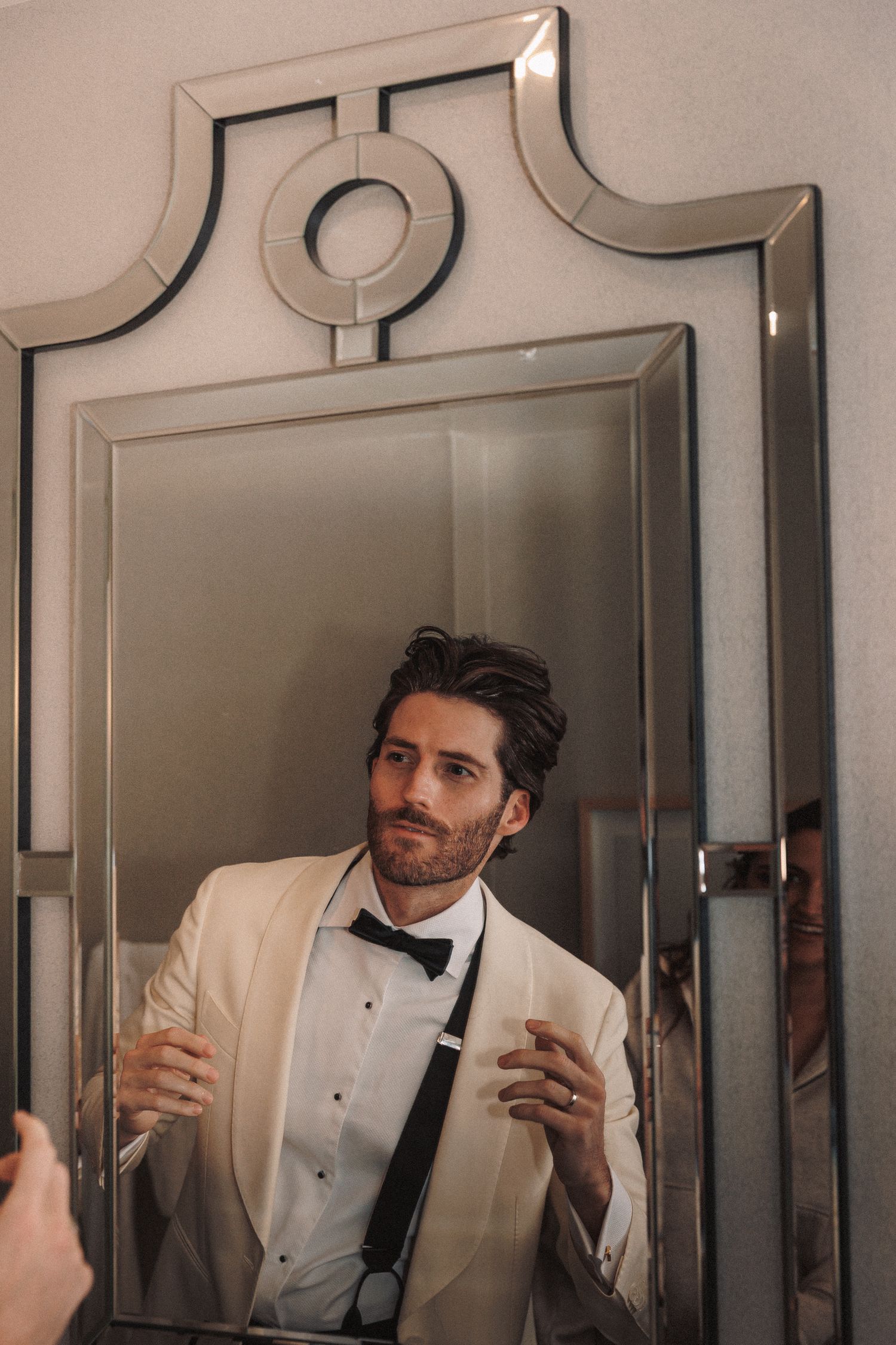 Groom in ivory tuxedo jacket and black bow tie checking his appearance in an ornate Art Deco mirror.