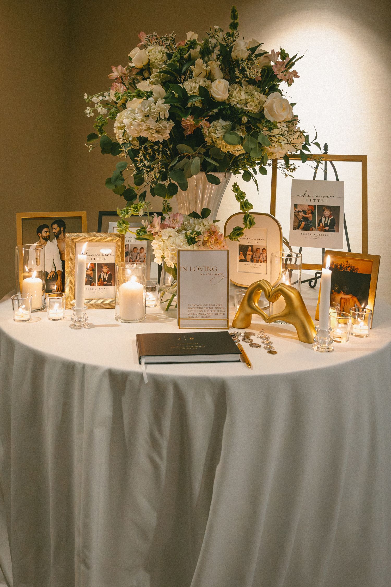 Wedding memory table with gold frames, candles, floral arrangement, and guest book on white linen.