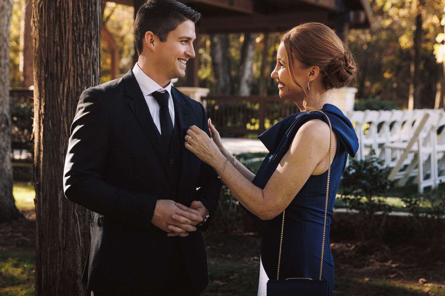 A woman in a blue gown adjusts a man's suit lapel outdoors at a wedding.