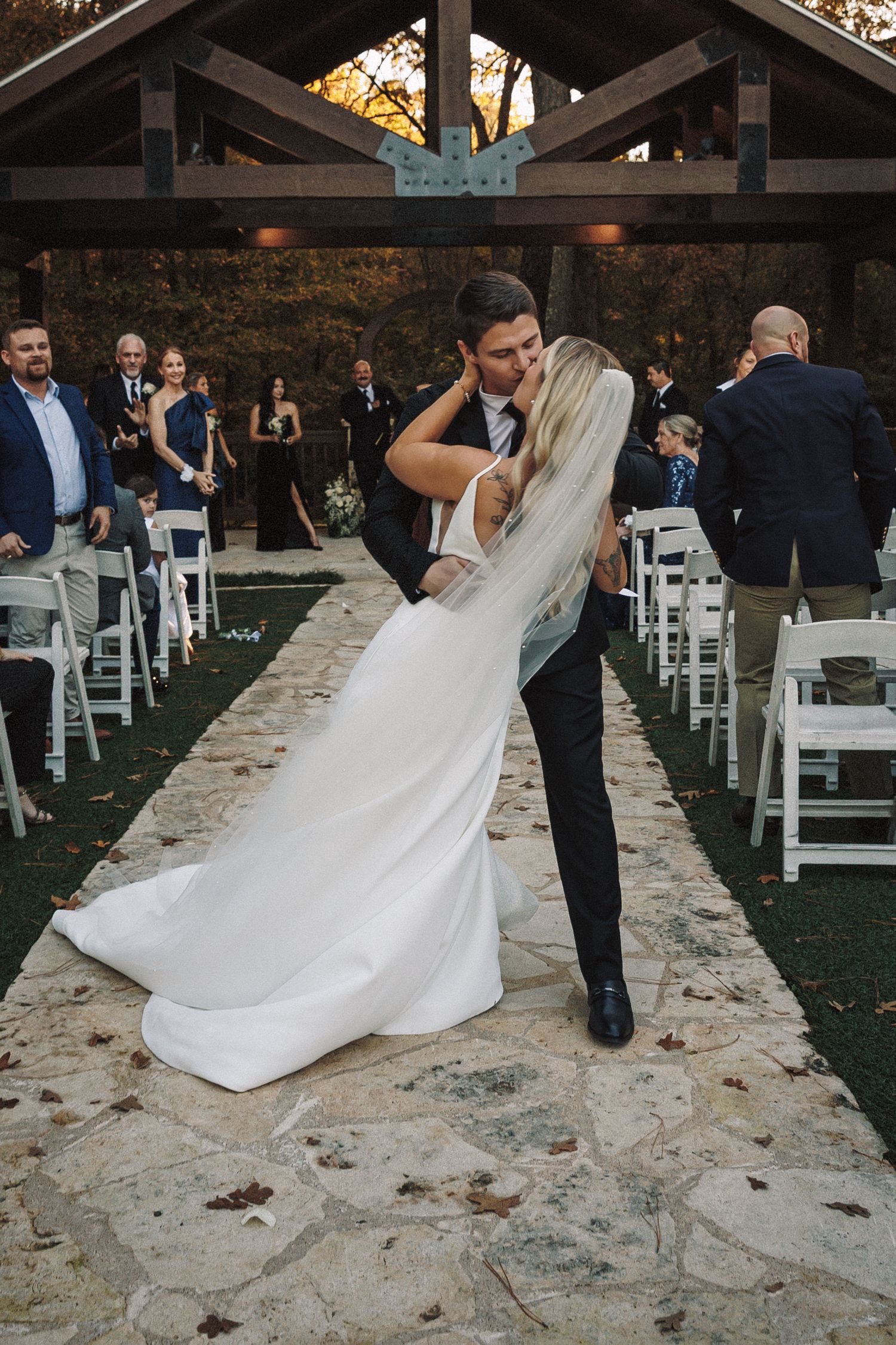 Groom dips bride for a romantic kiss in the stone aisle of an outdoor fall wedding ceremony pavilion.