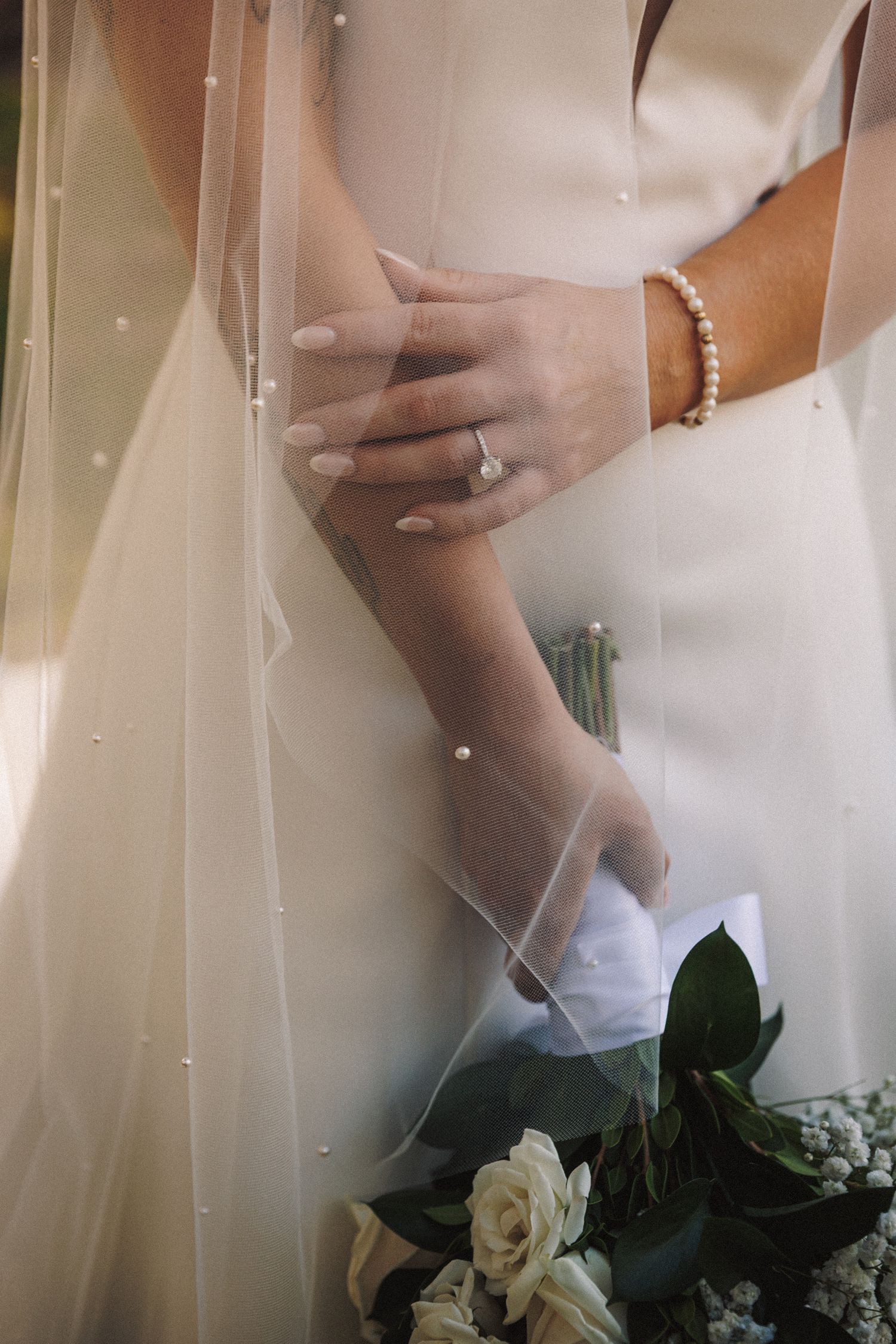 Close-up of bride's hands showing diamond engagement ring and pearl bracelet with white rose bouquet.
