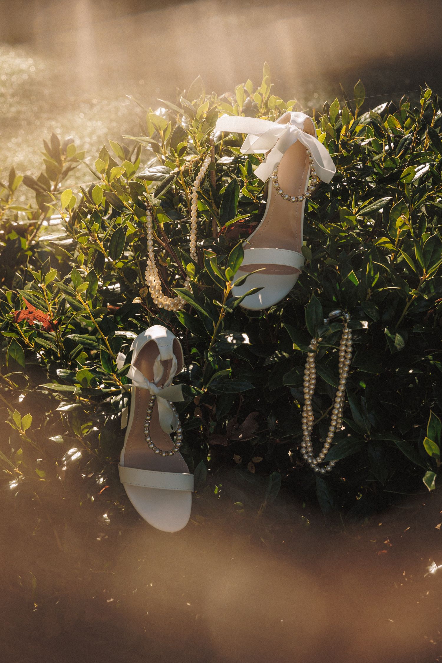 White bridal sandals with pearl necklaces draped over green bushes in golden sunlight.