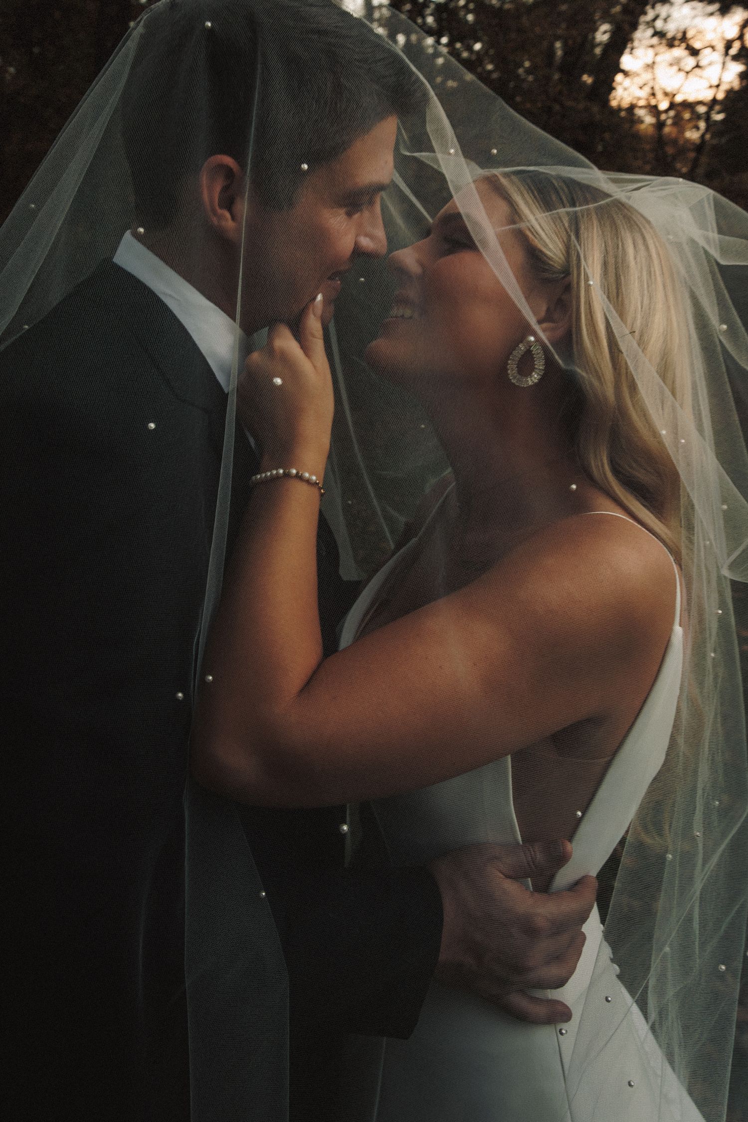 Bride and groom share a romantic moment under a pearl-adorned veil in warm golden light.