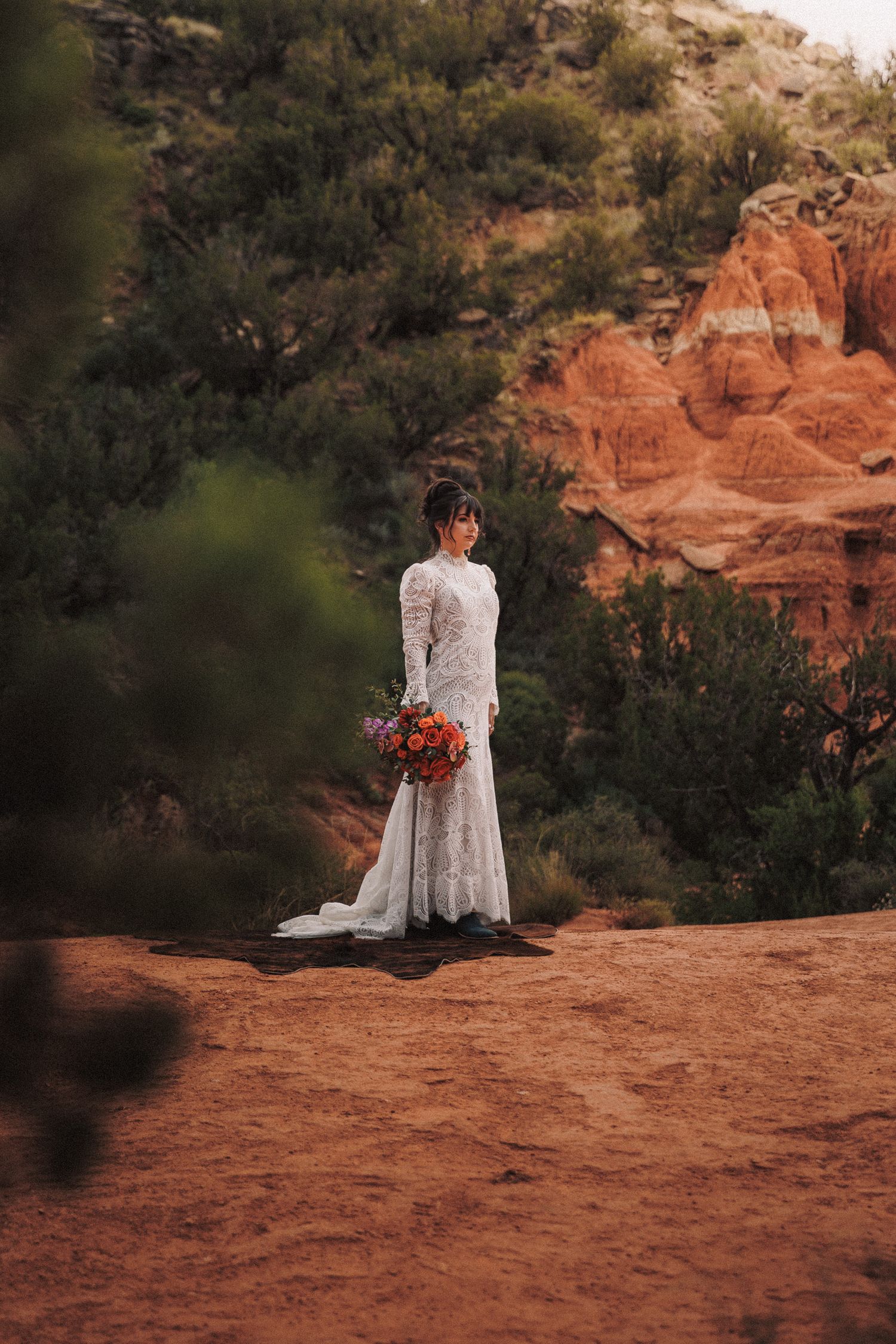 Bride in lace gown holding colorful bouquet stands on red rock formation with desert landscape in background.